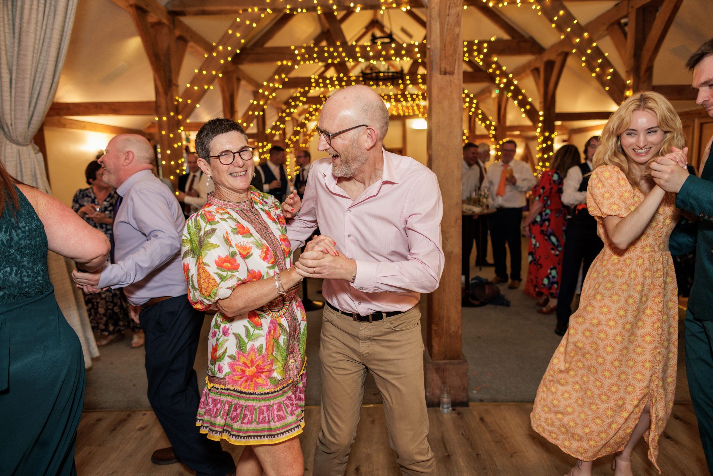 Couples dancing joyfully at a wedding reception in a rustic barn venue with warm fairy lights.