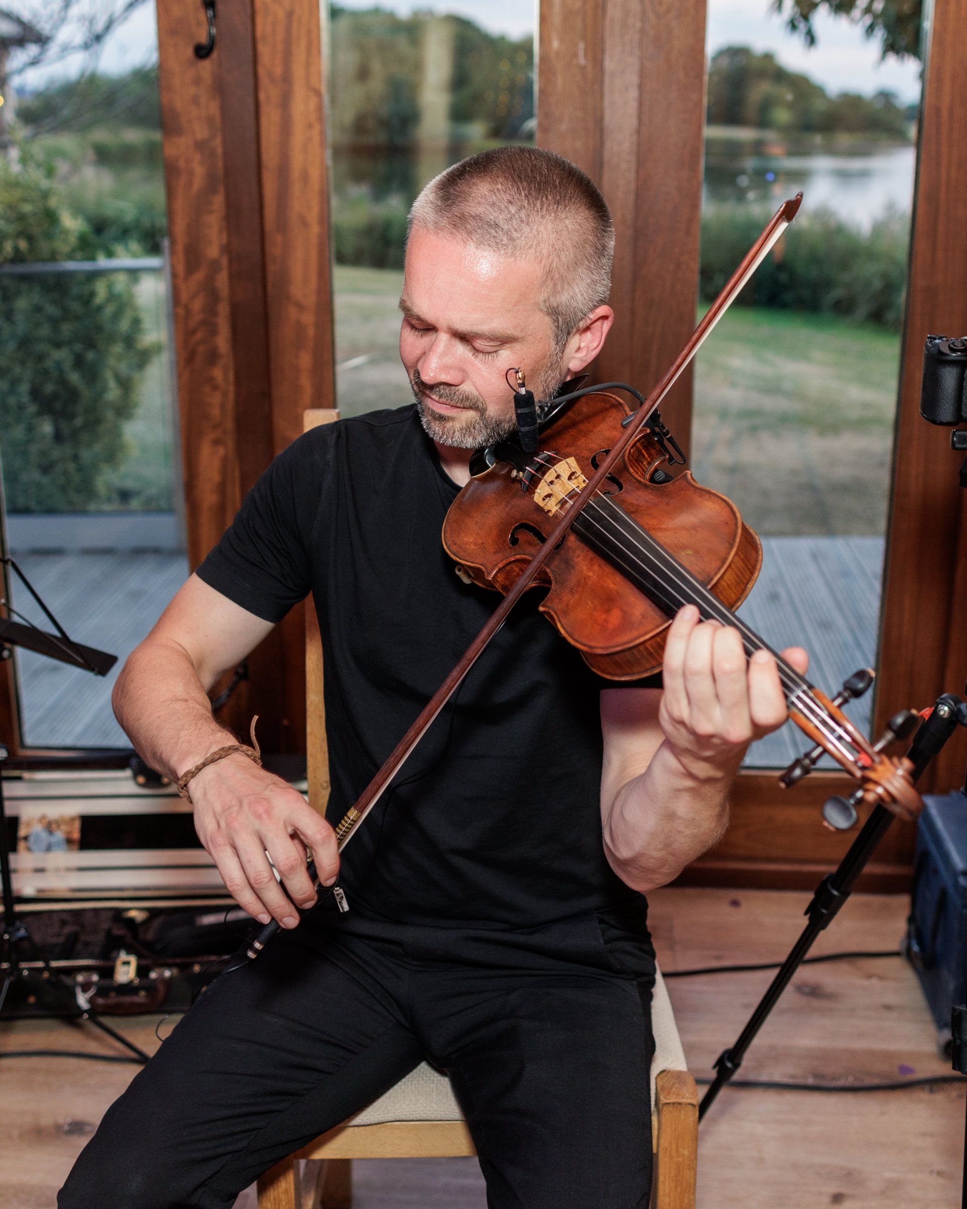 Man in black outfit playing violin indoors near large windows with scenic outdoor view.