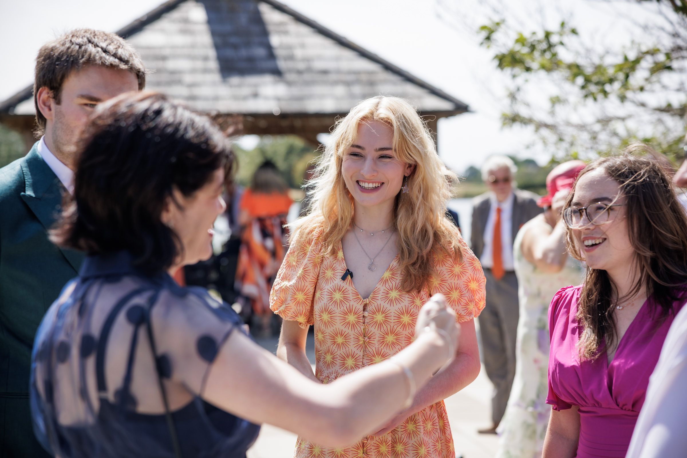 People socializing outdoors at a sunny event, woman in orange floral dress shaking hands and smiling.