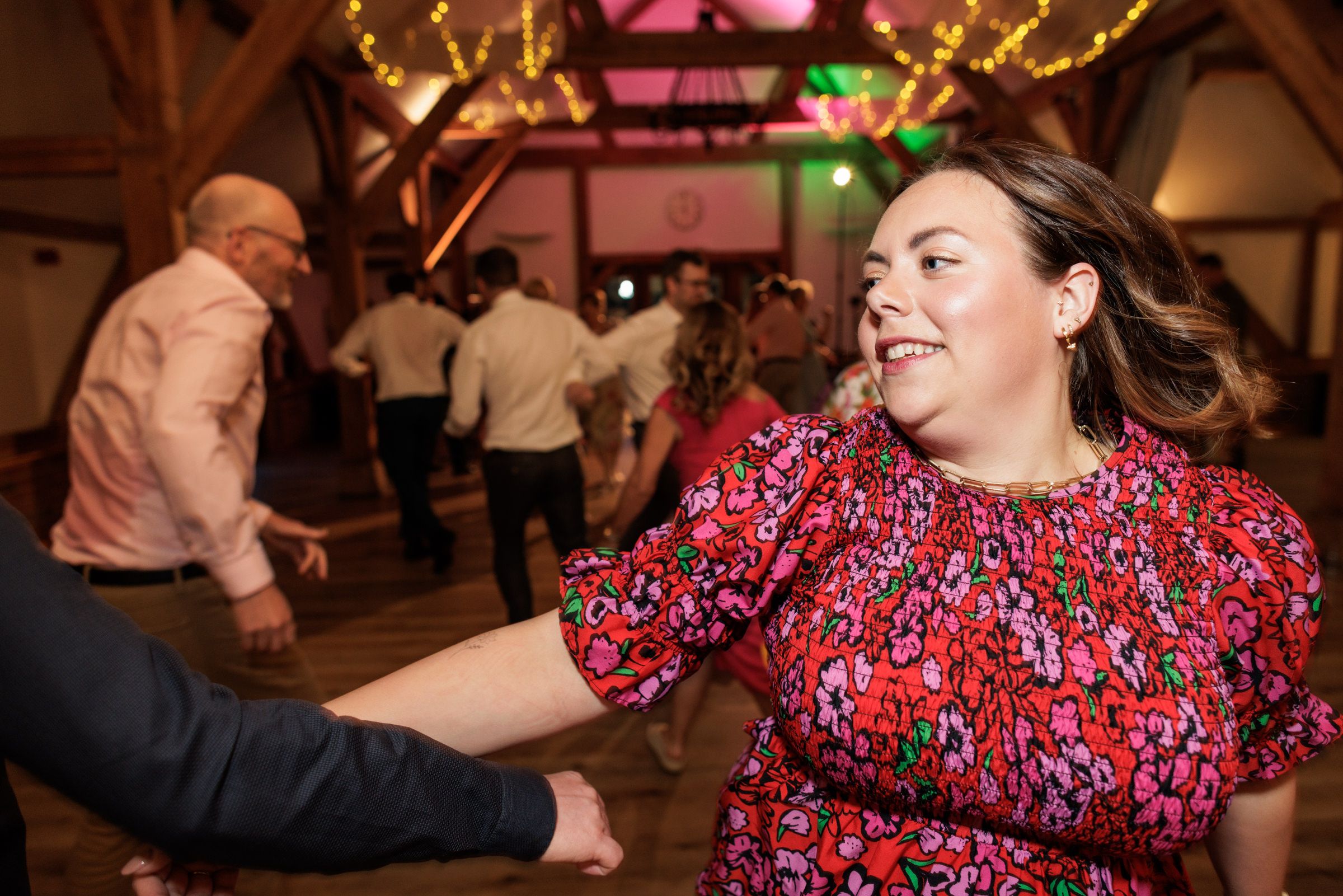 Woman in floral red dress dancing joyfully at a festive barn wedding reception with guests in background.