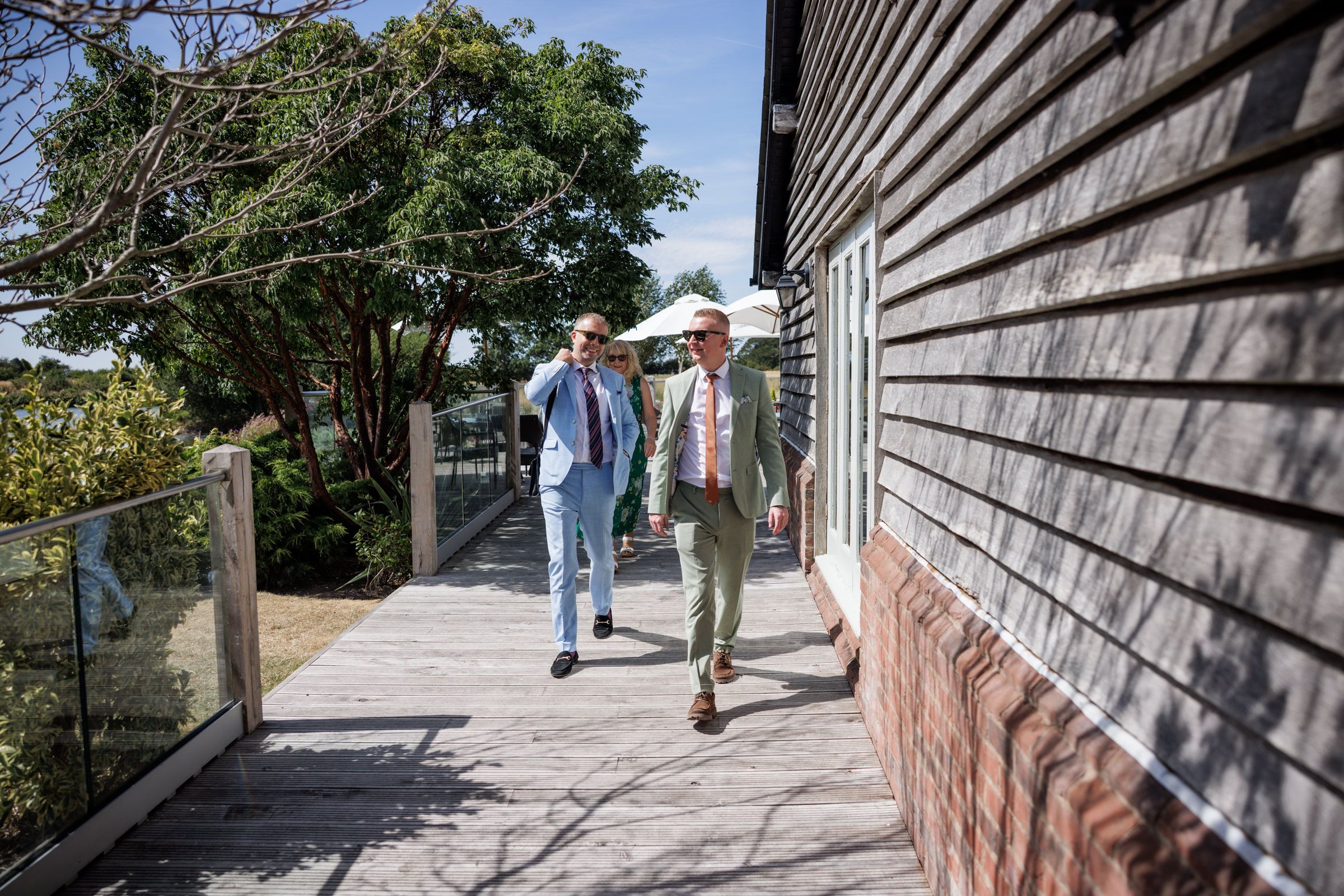 Two men in smart casual attire walk along a wooden deck beside a dark timber building on a sunny day.
