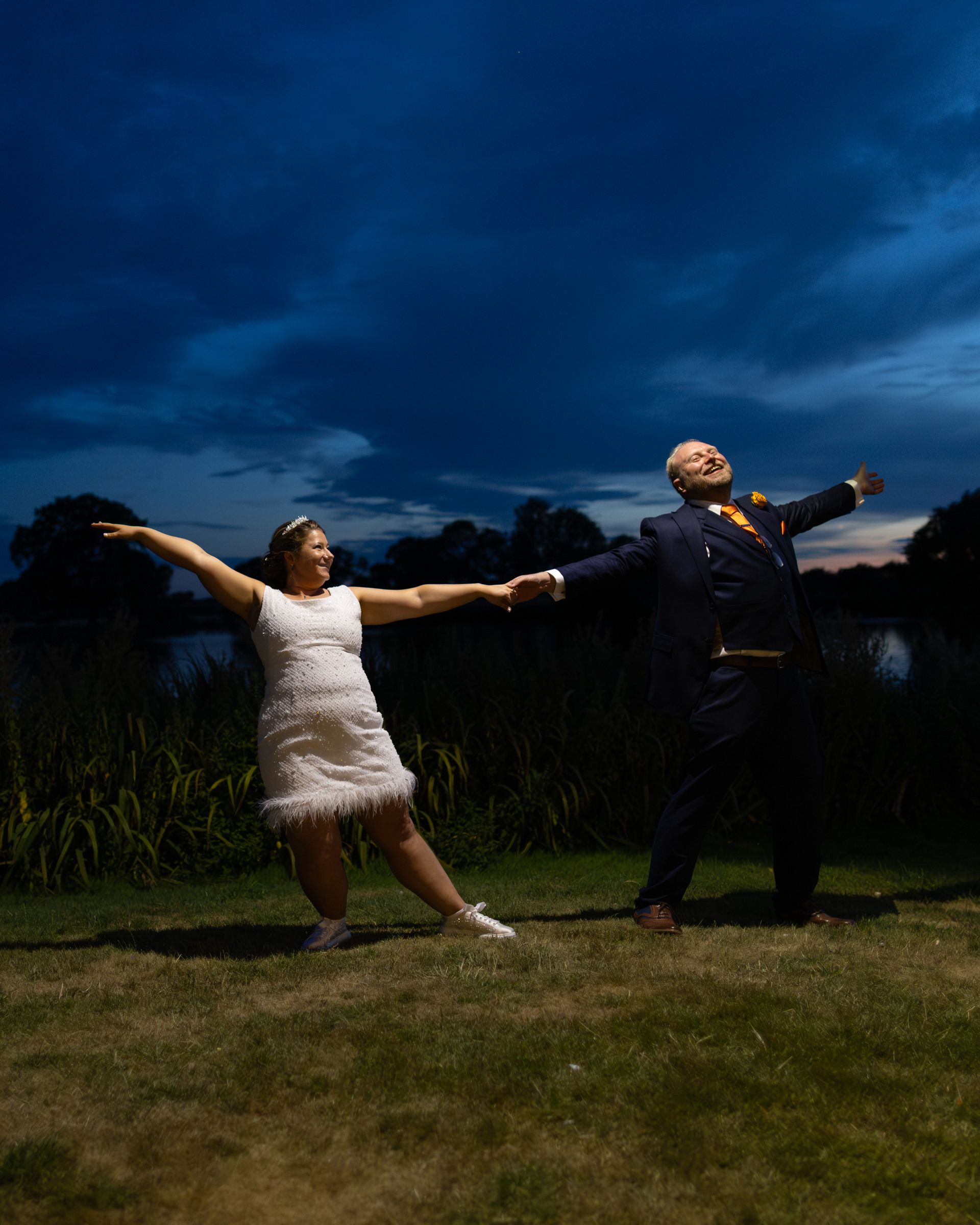 Joyful couple posing dramatically at dusk, arms outstretched, in wedding attire on a lush lawn under a moody blue sky.