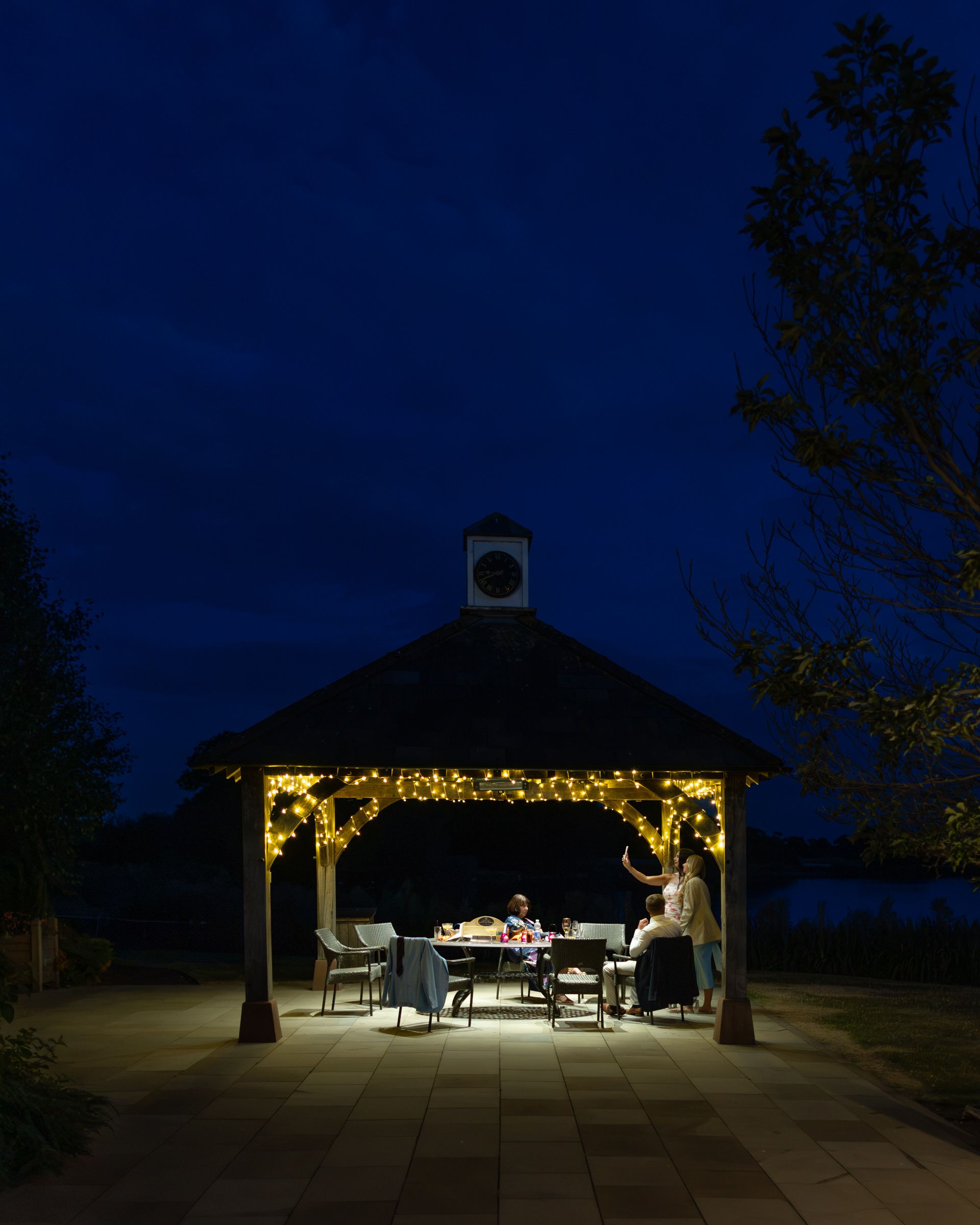 People dining under a illuminated gazebo with fairy lights at dusk in a serene outdoor setting.