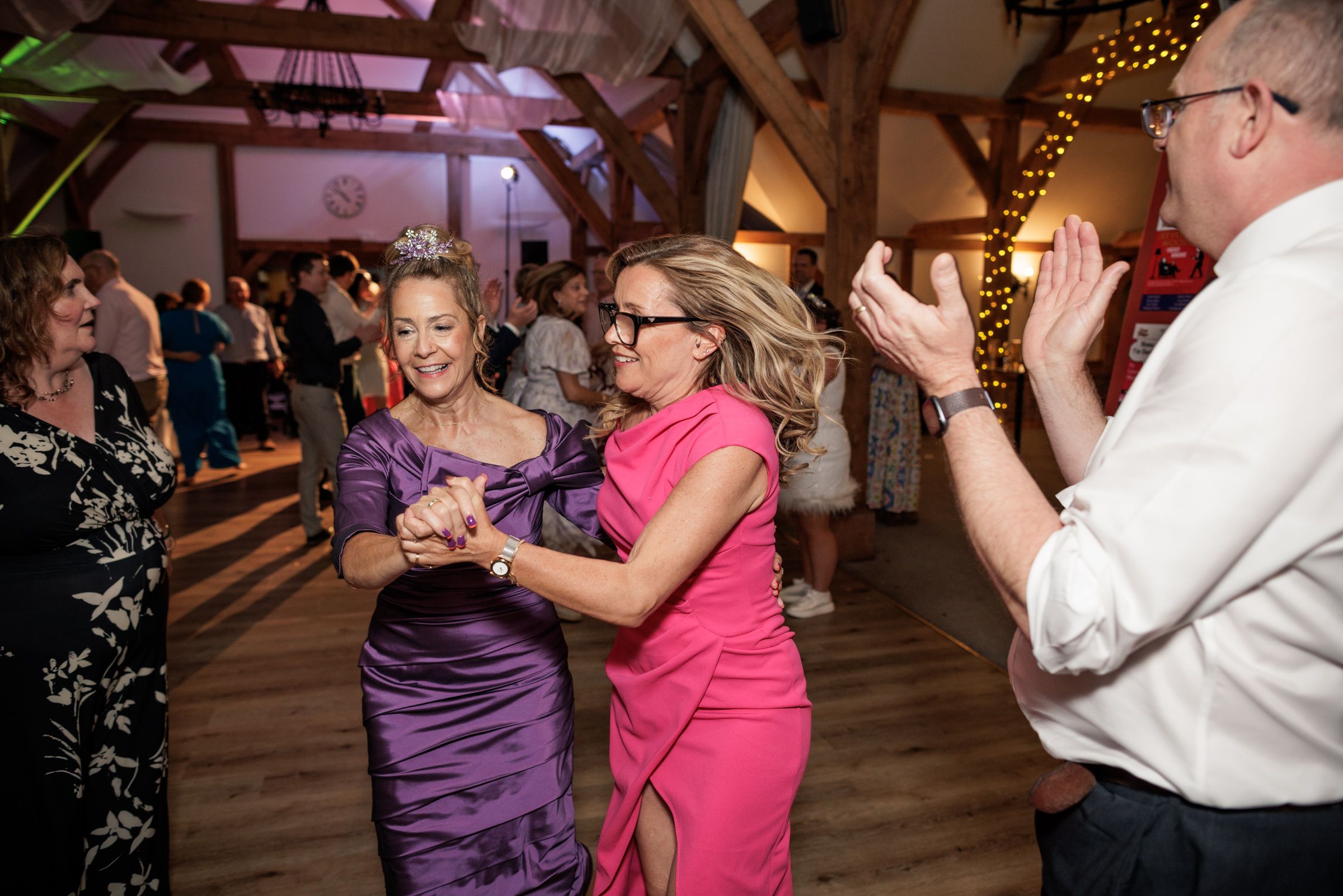 Two women in elegant dresses dancing joyfully at a wedding reception in a rustic venue with warm lighting.
