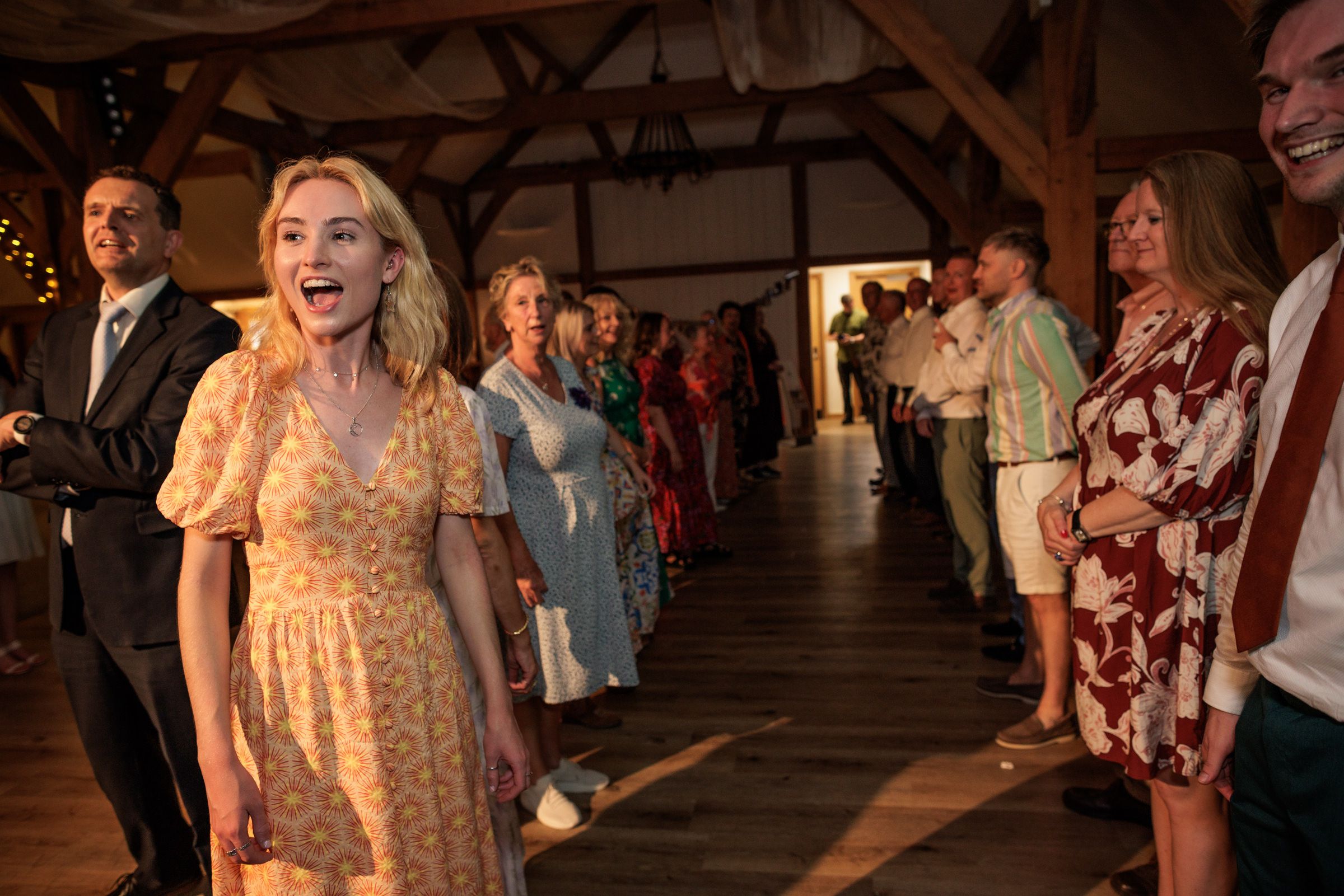 Guests in colorful attire laugh and dance at a rustic barn wedding reception with warm lighting.