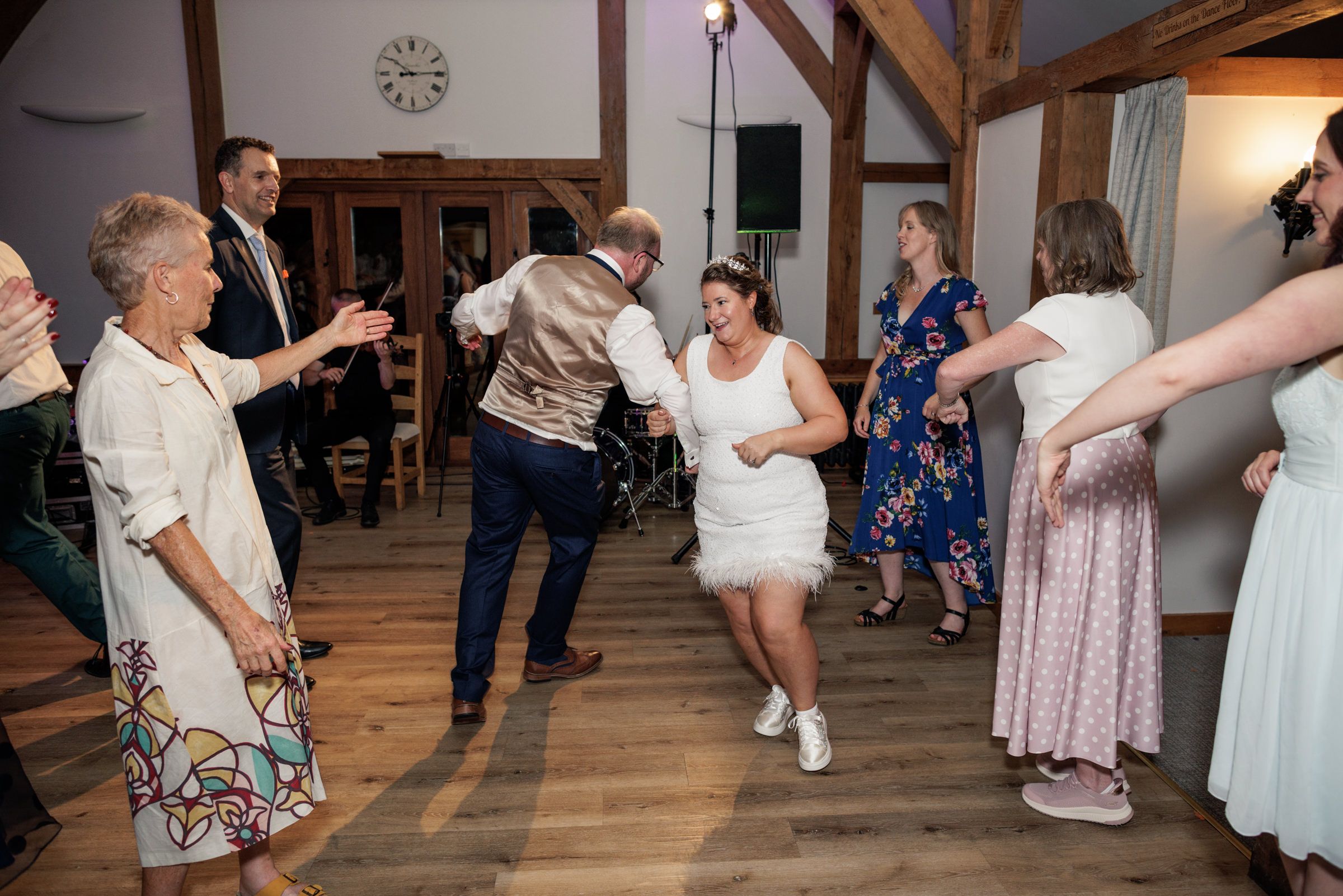 Guests dancing energetically at a wedding reception in a rustic barn venue, with bride in white dress joining the celebration.