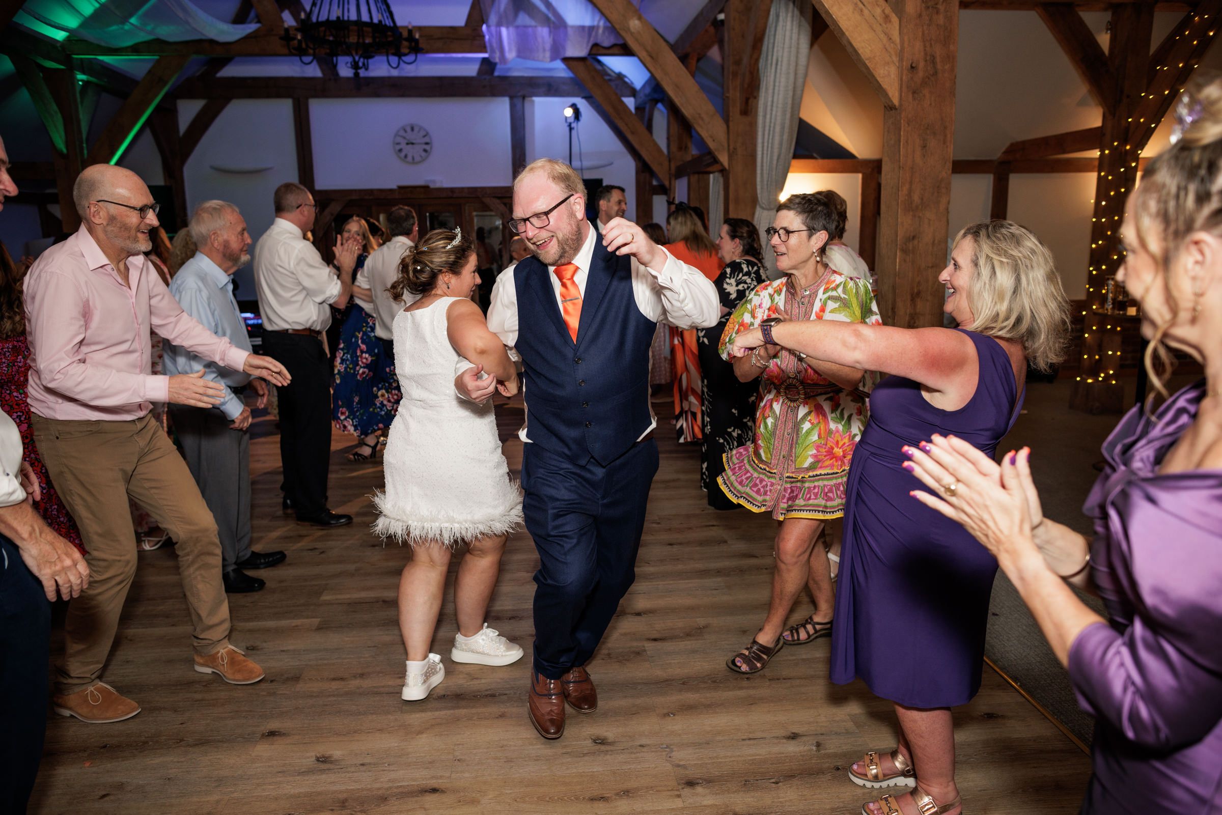 Guests dancing joyfully at a rustic barn wedding reception with string lights and wooden beams.
