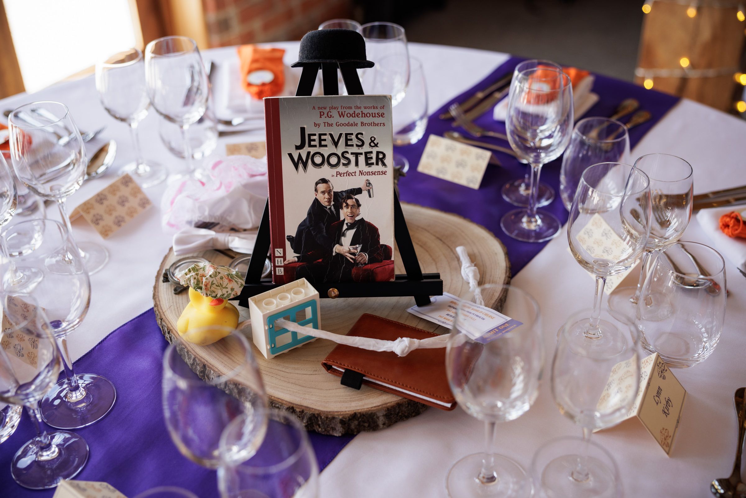Elegant wedding table setting with Jeeves & Wooster book centerpiece, purple linens, crystal glasses, and decorative wood slice.