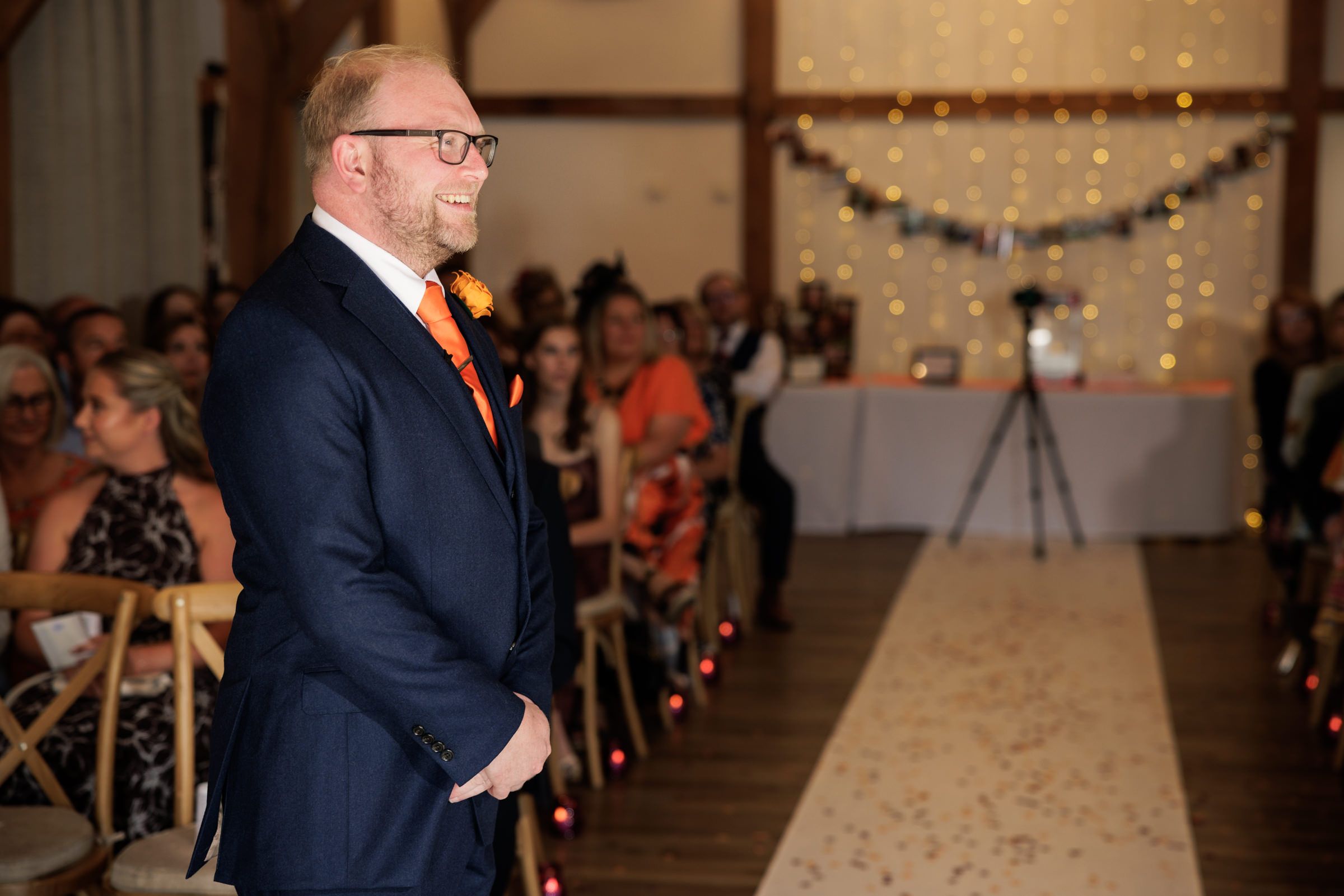 Groom in navy suit with orange tie stands at altar awaiting bride at elegant barn wedding ceremony.