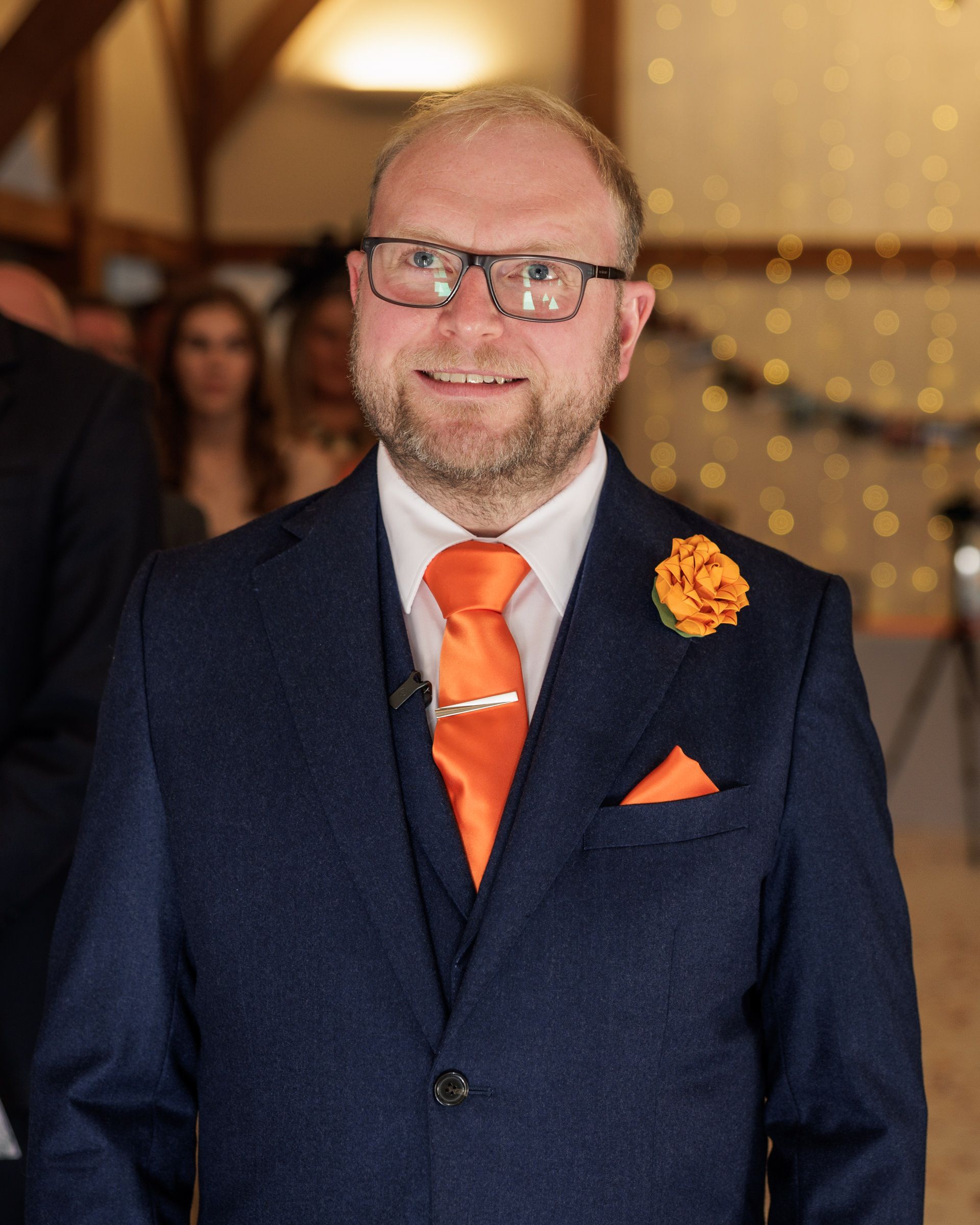 Man in navy suit with orange tie, boutonniere, and pocket square smiling at formal event.