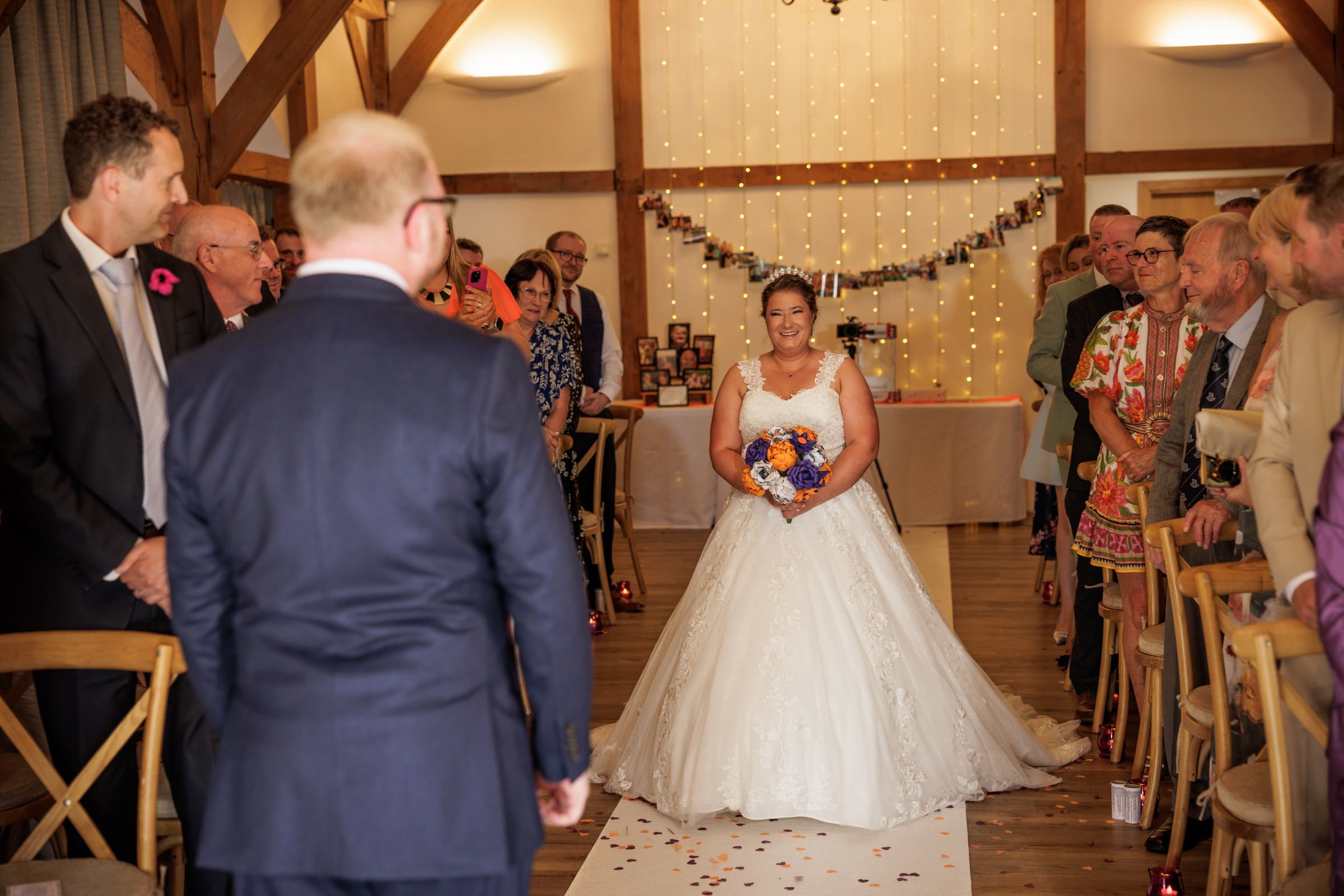 Bride in white ball gown walks down the aisle holding colorful bouquet at an elegant indoor wedding ceremony.