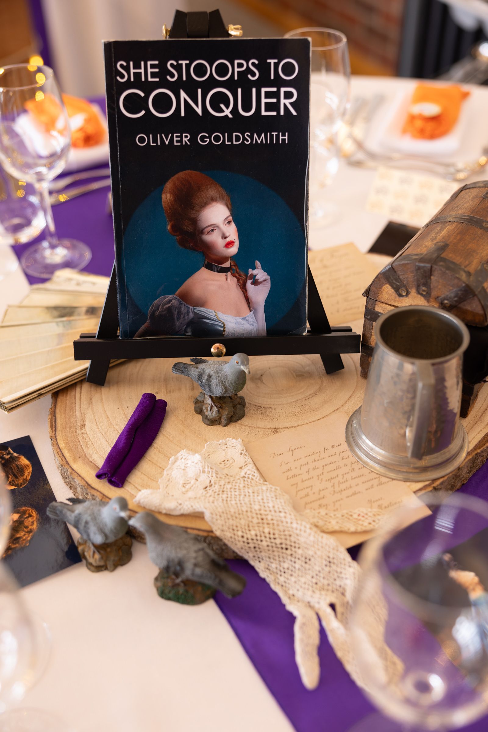 Table centerpiece featuring She Stoops to Conquer by Oliver Goldsmith on easel with vintage props and purple accents.
