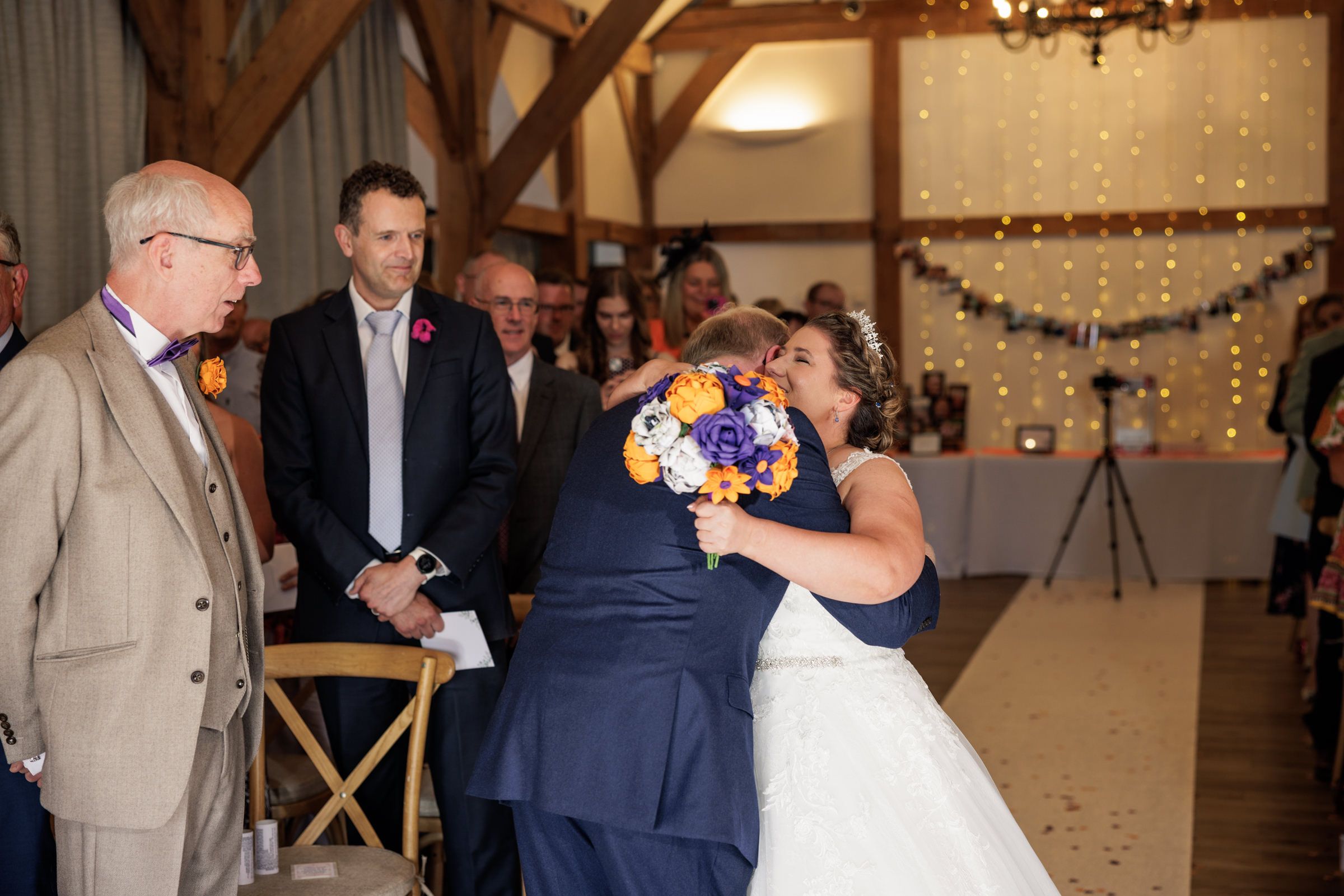 Bride and groom share a heartfelt hug during their wedding ceremony in a rustic barn venue with fairy lights.