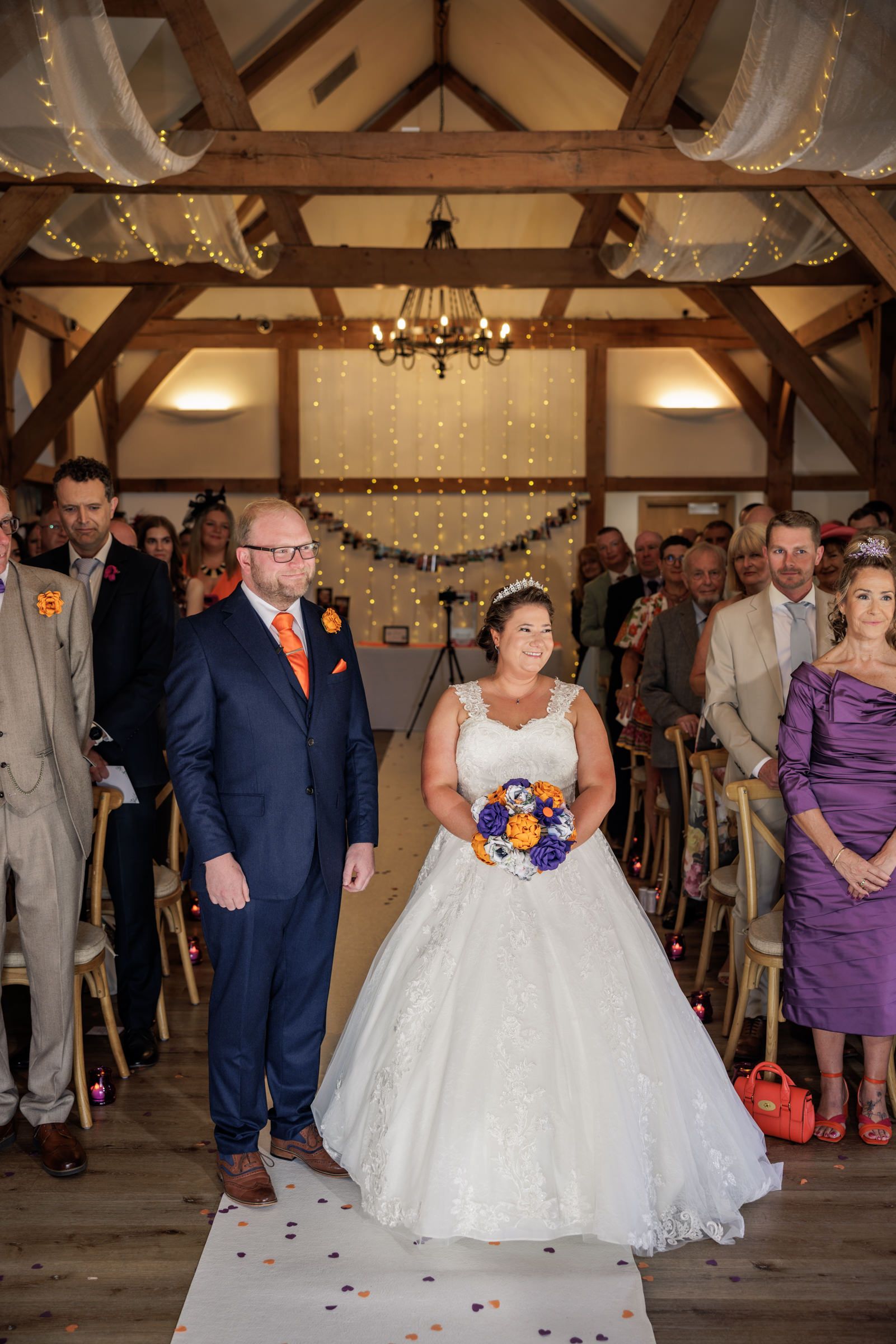 Bride and groom walk down the aisle at a rustic wedding venue decorated with fairy lights and wooden beams.