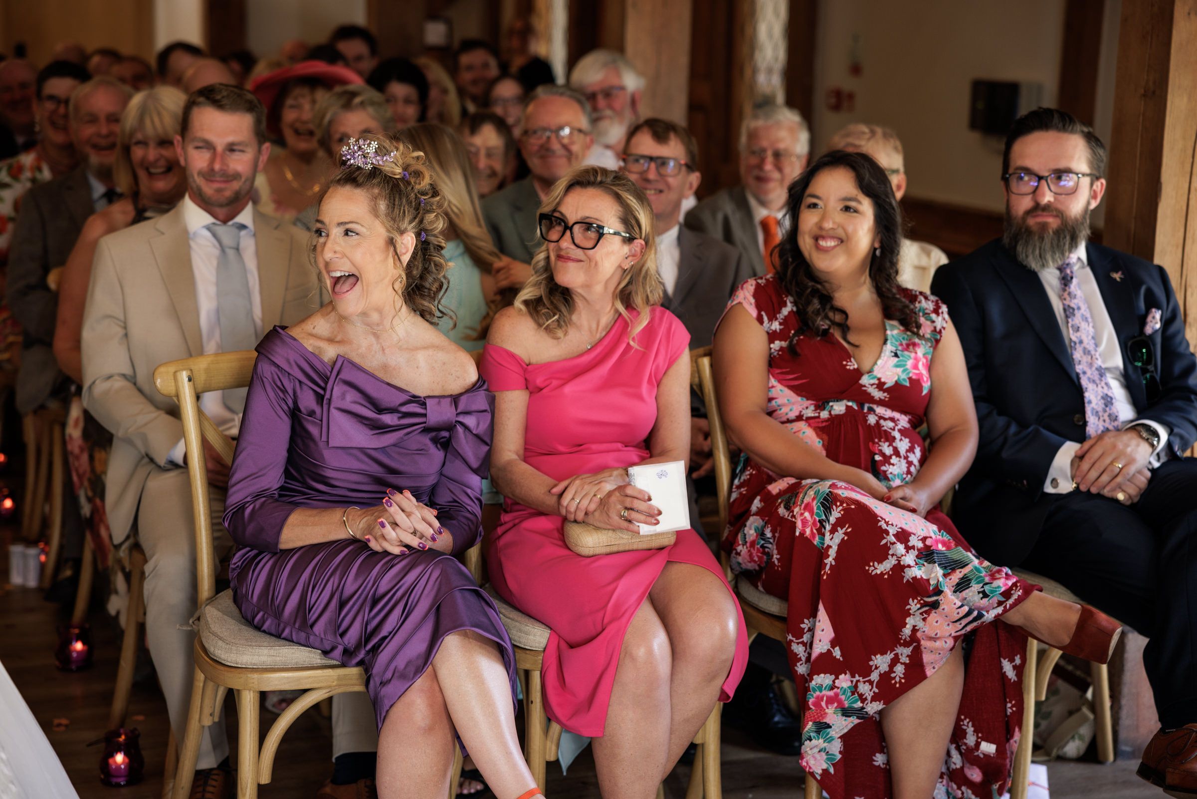 Guests laughing and smiling at a wedding ceremony, seated in rows wearing elegant formal attire.