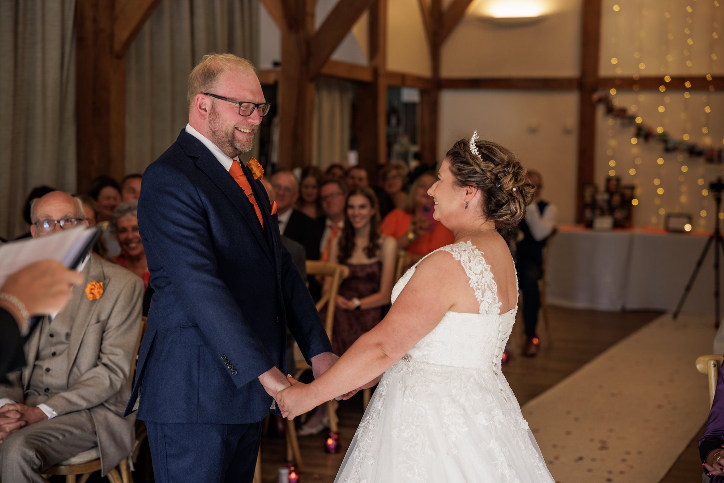 Bride and groom holding hands and smiling during indoor wedding ceremony with guests seated in background.