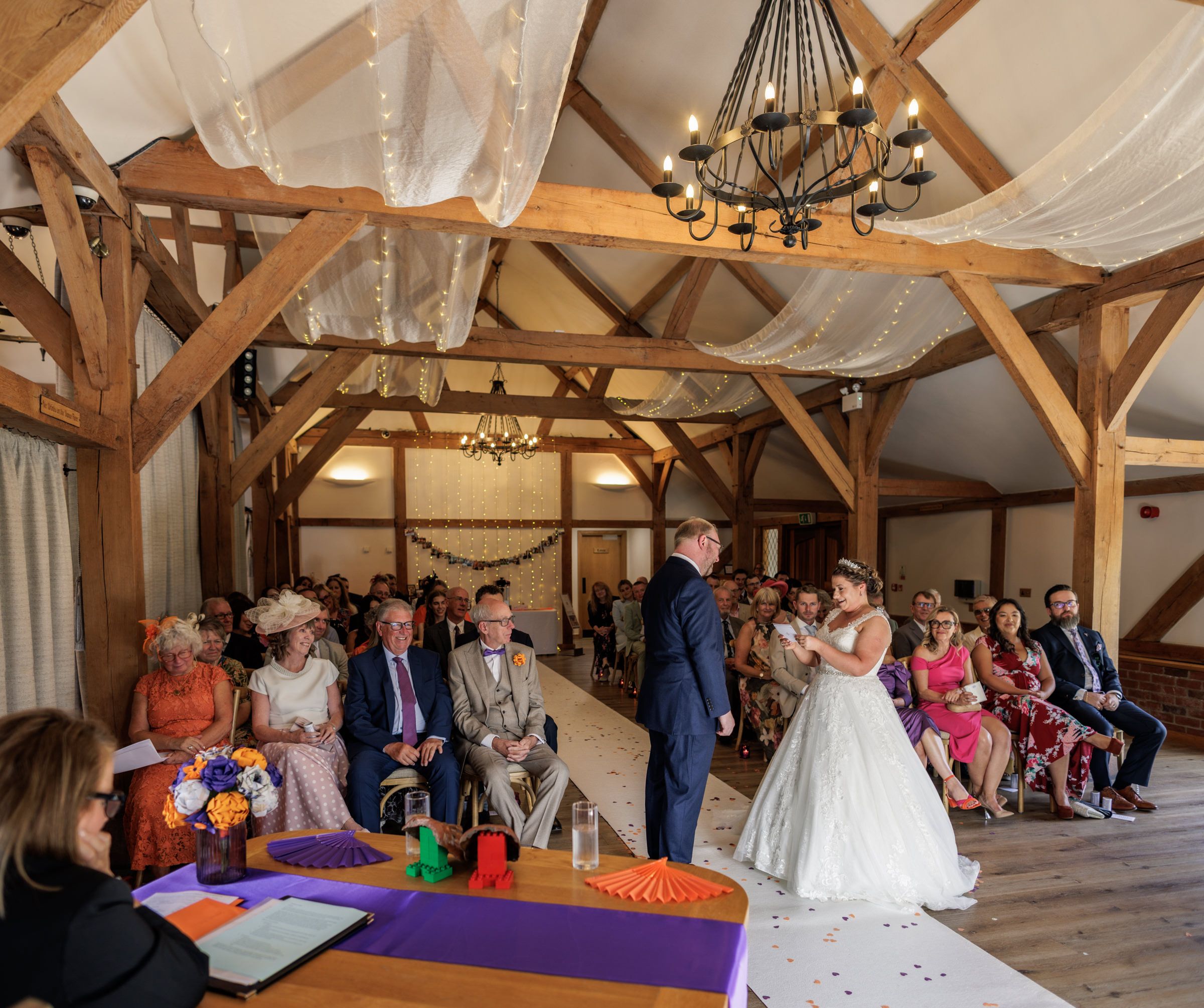 Bride and groom exchange vows in a rustic barn wedding venue with wooden beams, draped fabric, and seated guests.