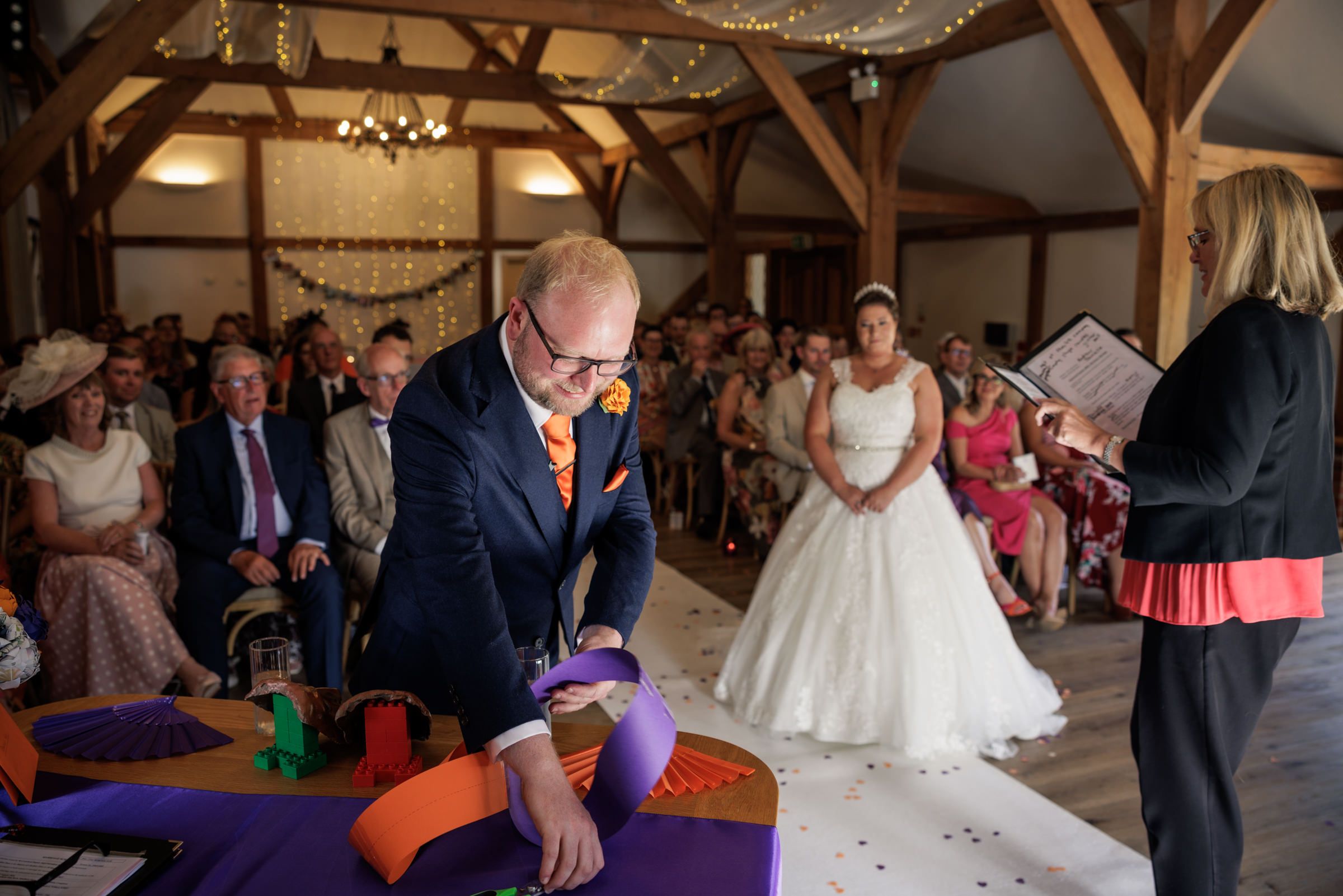 Groom in navy suit performs sand ceremony at rustic barn wedding while bride in white gown watches with guests seated behind.
