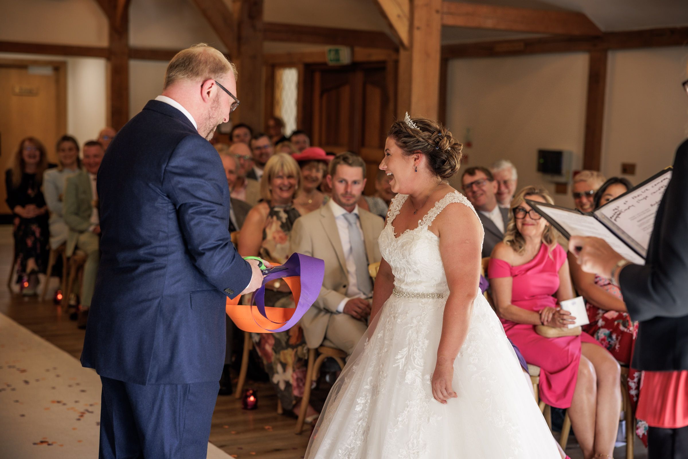 Groom in navy suit presents bride in white lace gown with a colorful gift during wedding ceremony indoors.