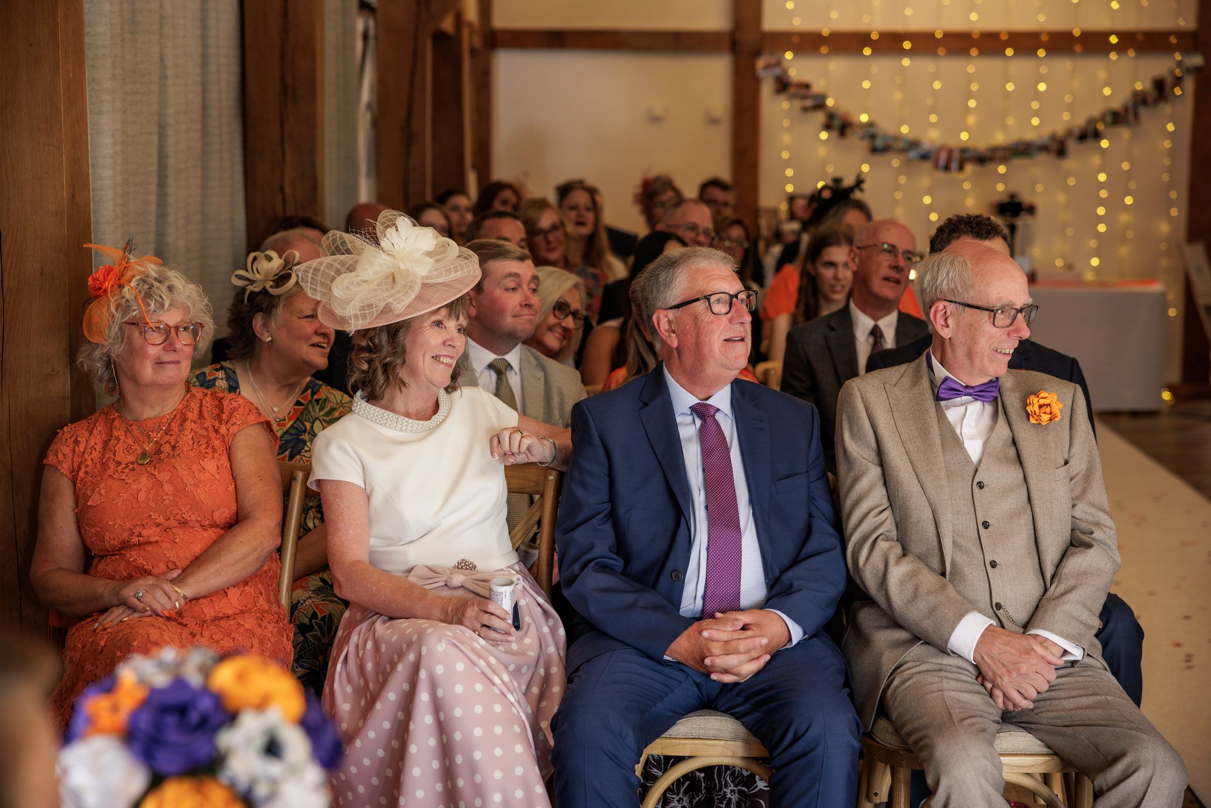 Guests smiling and laughing during a colorful wedding ceremony, seated in rows wearing formal attire and festive hats.