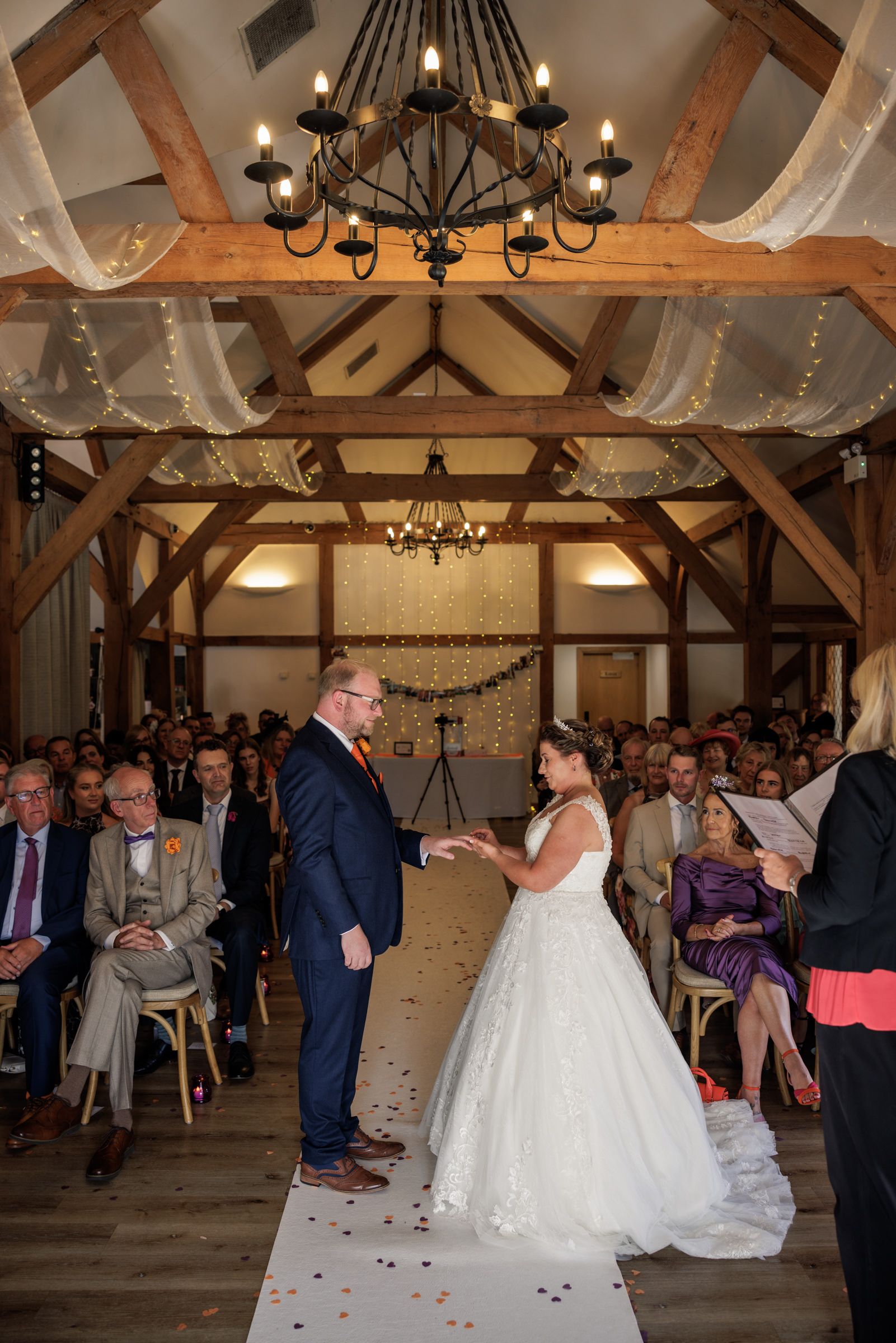 Bride and groom exchange vows in a decorated barn wedding venue with wooden beams, fairy lights, and seated guests.