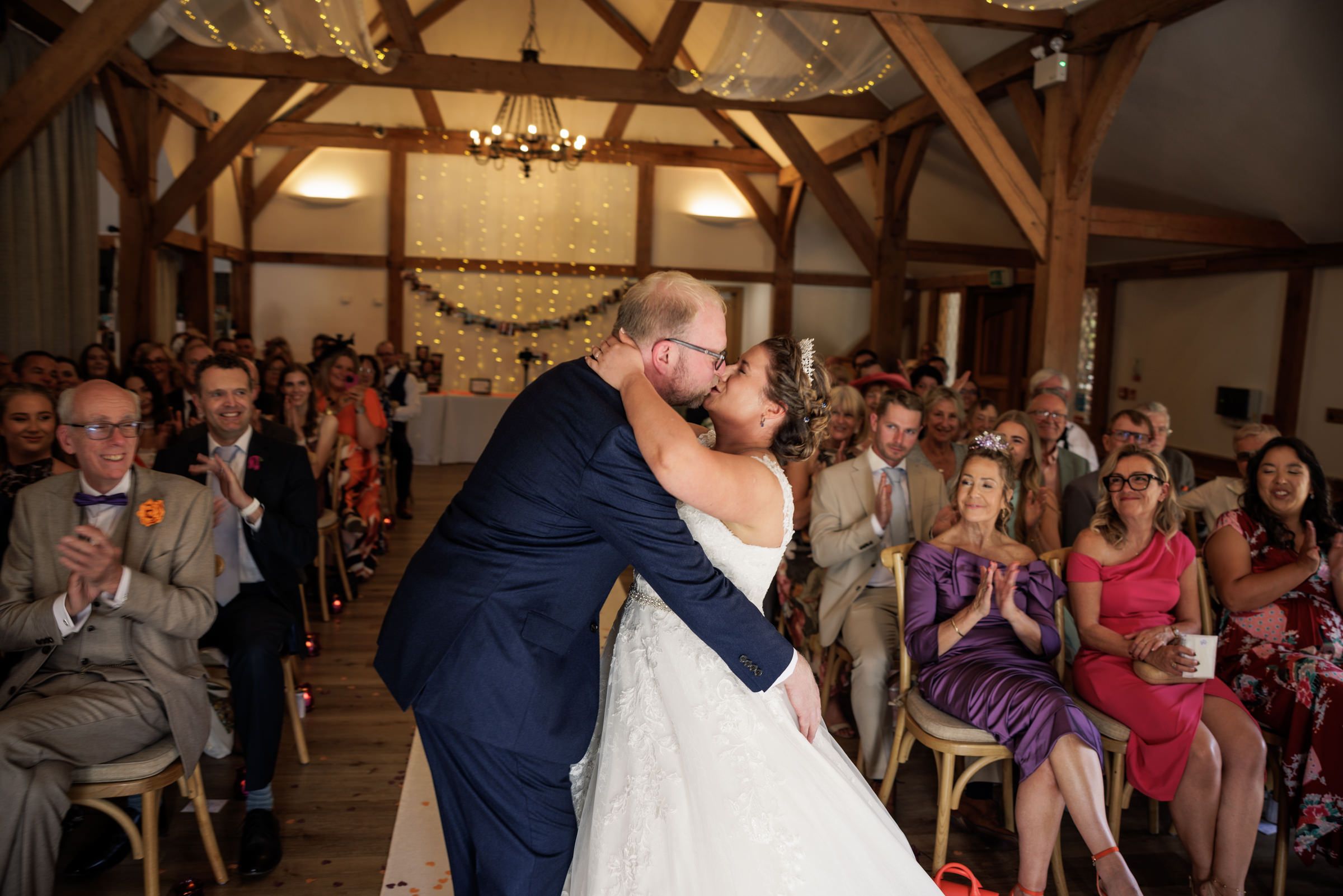 A bride and groom share their first kiss at a rustic barn wedding ceremony while guests applaud in the background.