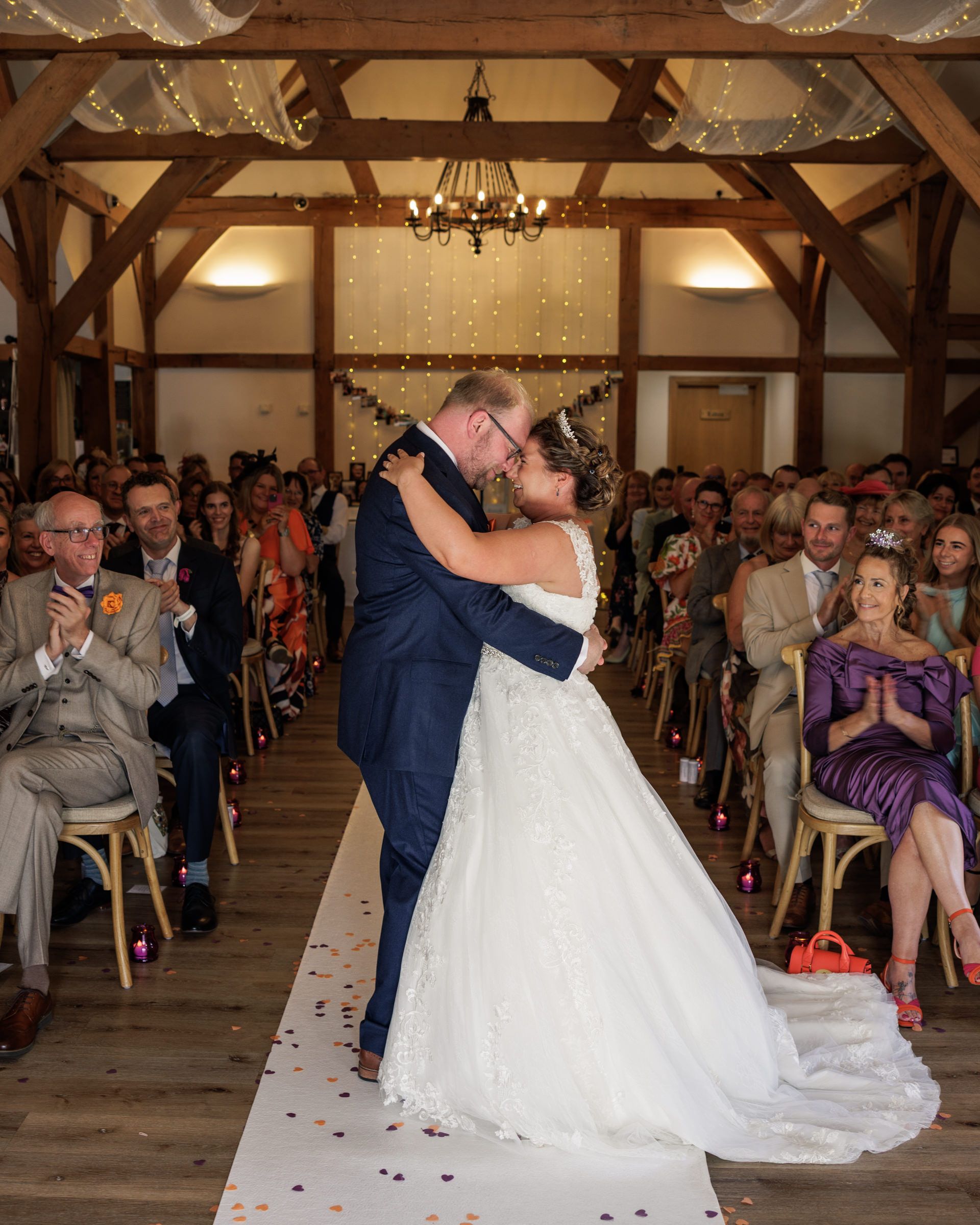 Bride and groom share first kiss at rustic barn wedding ceremony with confetti and guests applauding in background.