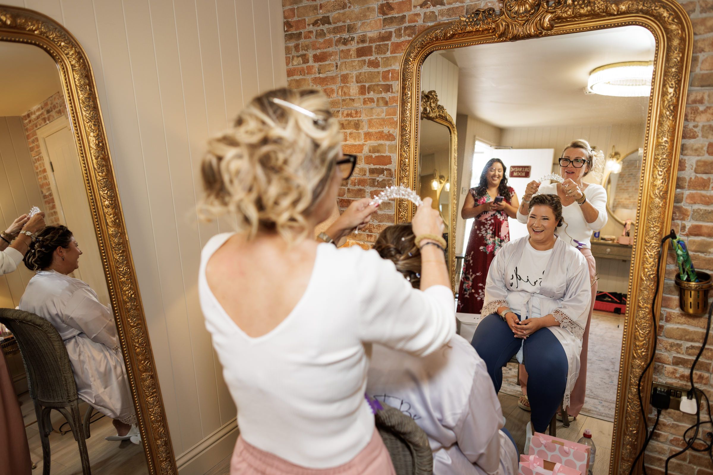 Bridal party getting ready in a rustic room with gold mirrors, hairstylist working on bride's updo.