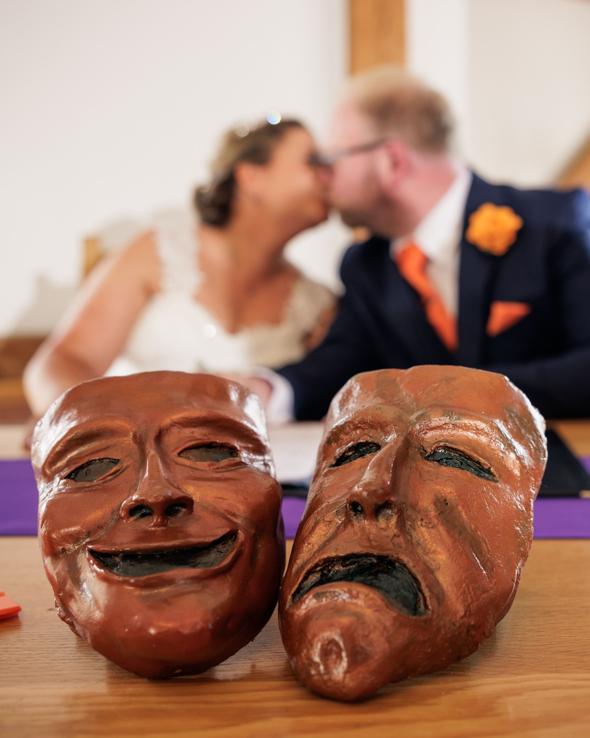 Comedy and tragedy masks in foreground with a bride and groom sharing a kiss at their wedding ceremony behind.