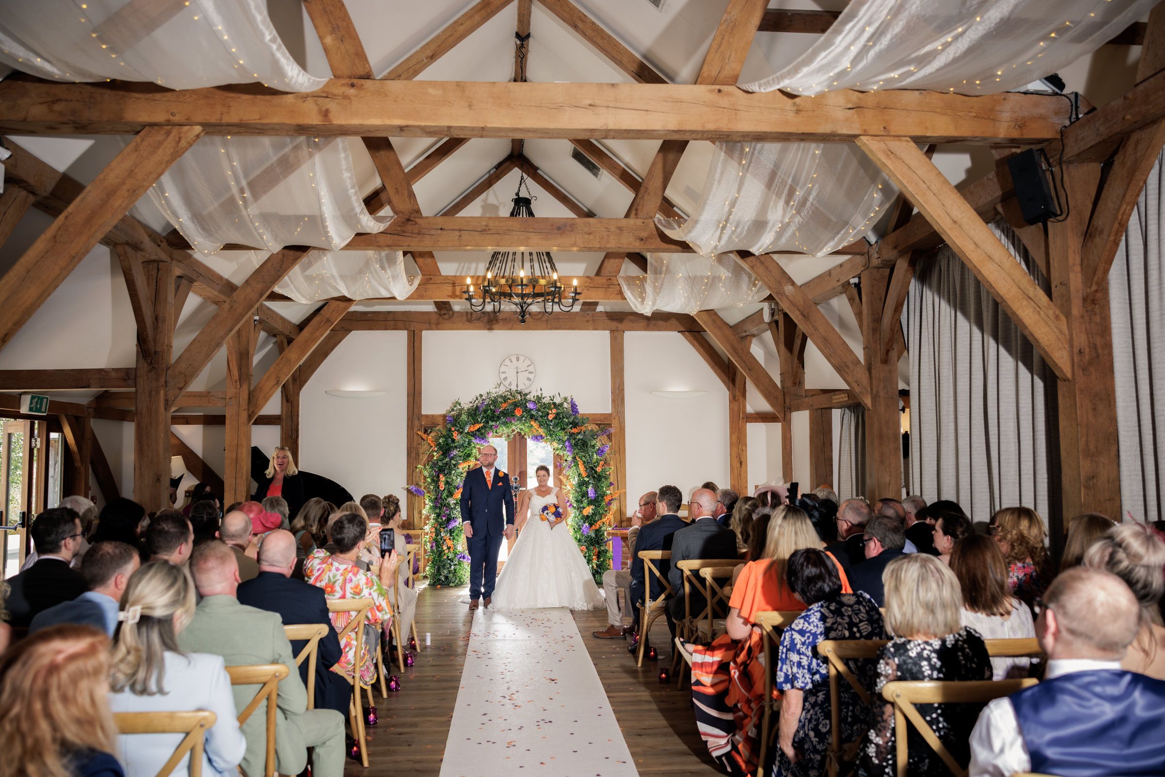 Elegant barn wedding ceremony with wooden beams, floral arch, and guests seated along a white petal aisle.