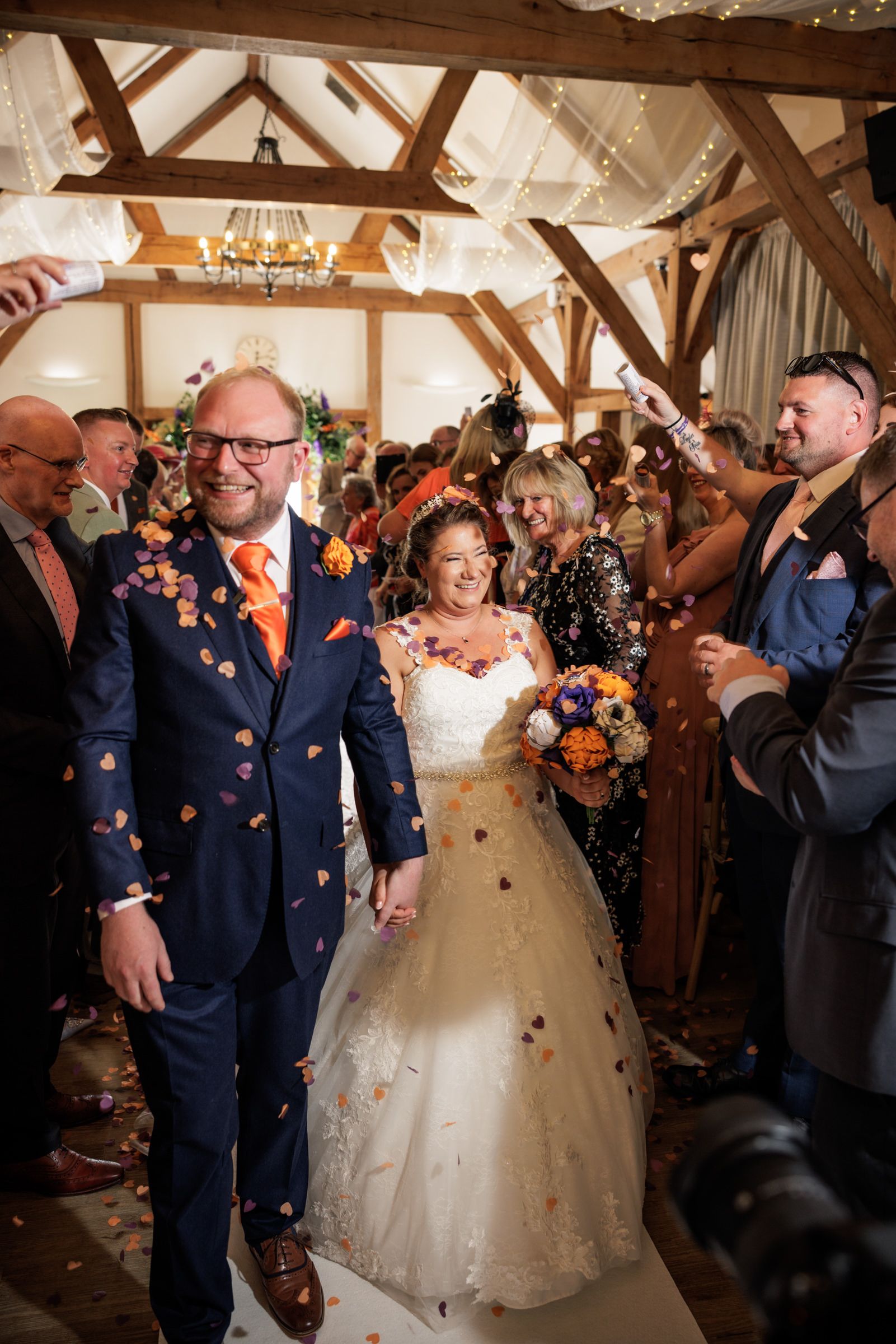Bride and groom walk through confetti shower in rustic barn venue, groom in navy suit with orange tie, bride in white gown.