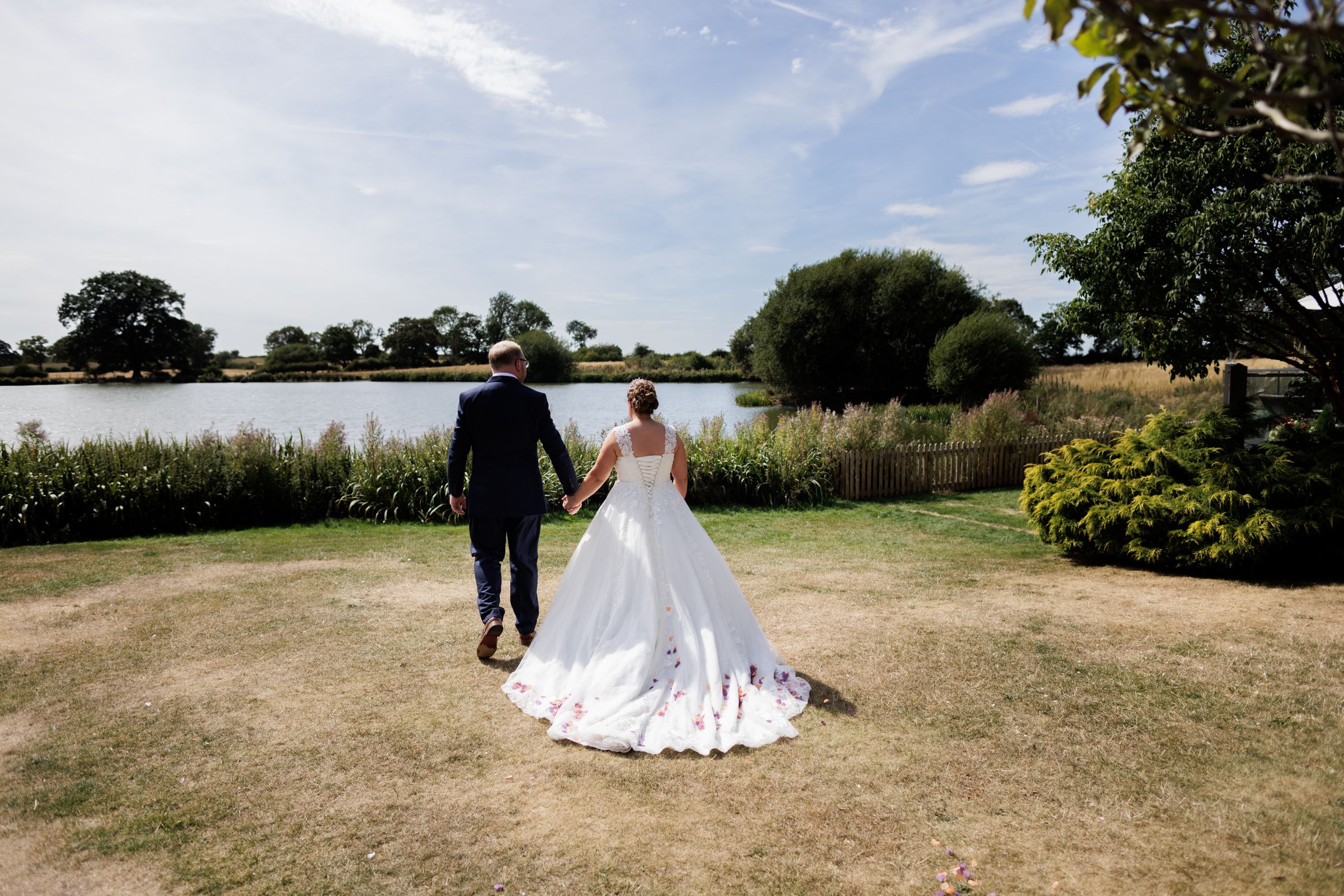Bride and groom holding hands walking towards a scenic lake on a sunny wedding day in a lush garden setting.