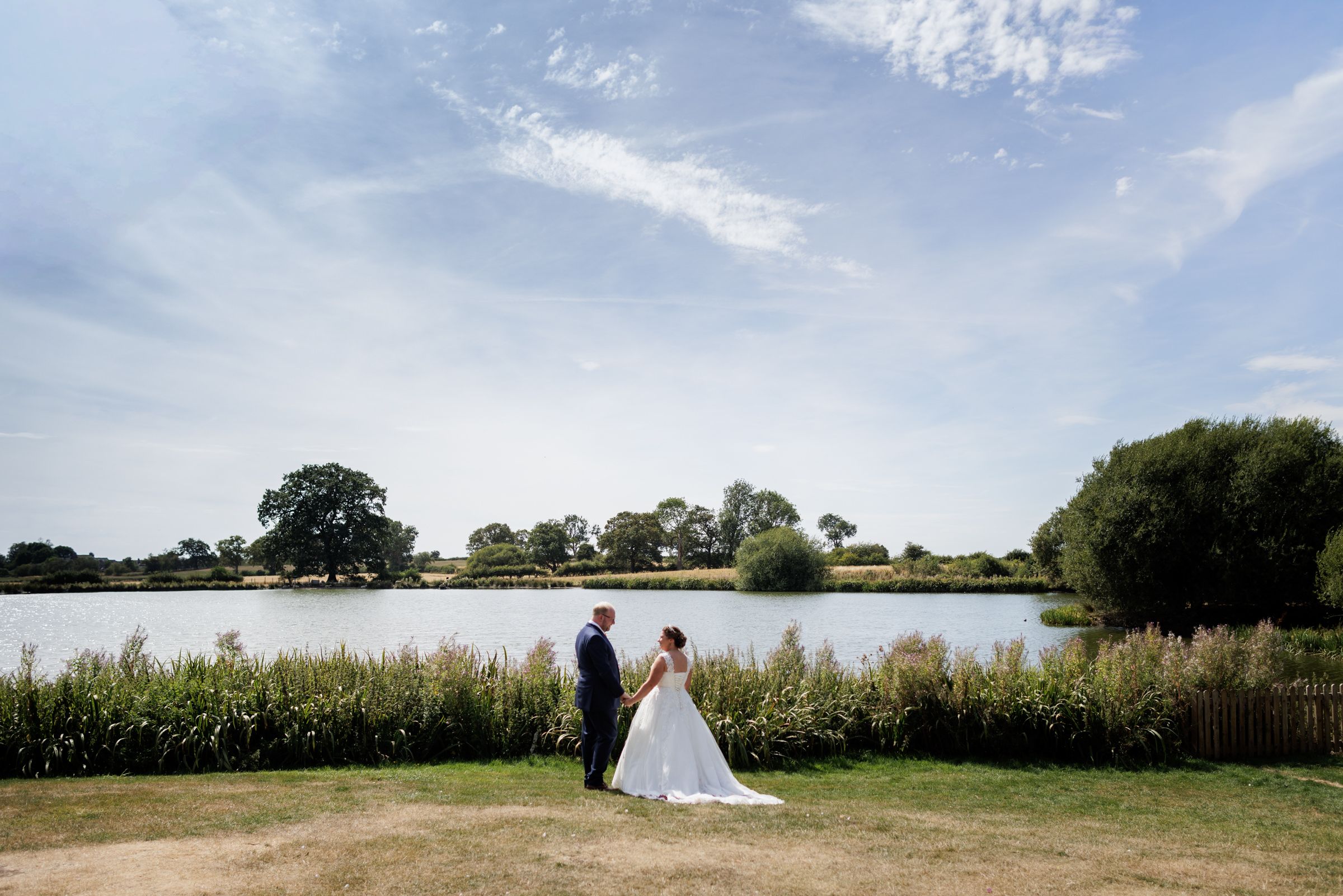 Bride and groom standing by a serene lake surrounded by lush greenery under a bright blue sky on their wedding day.