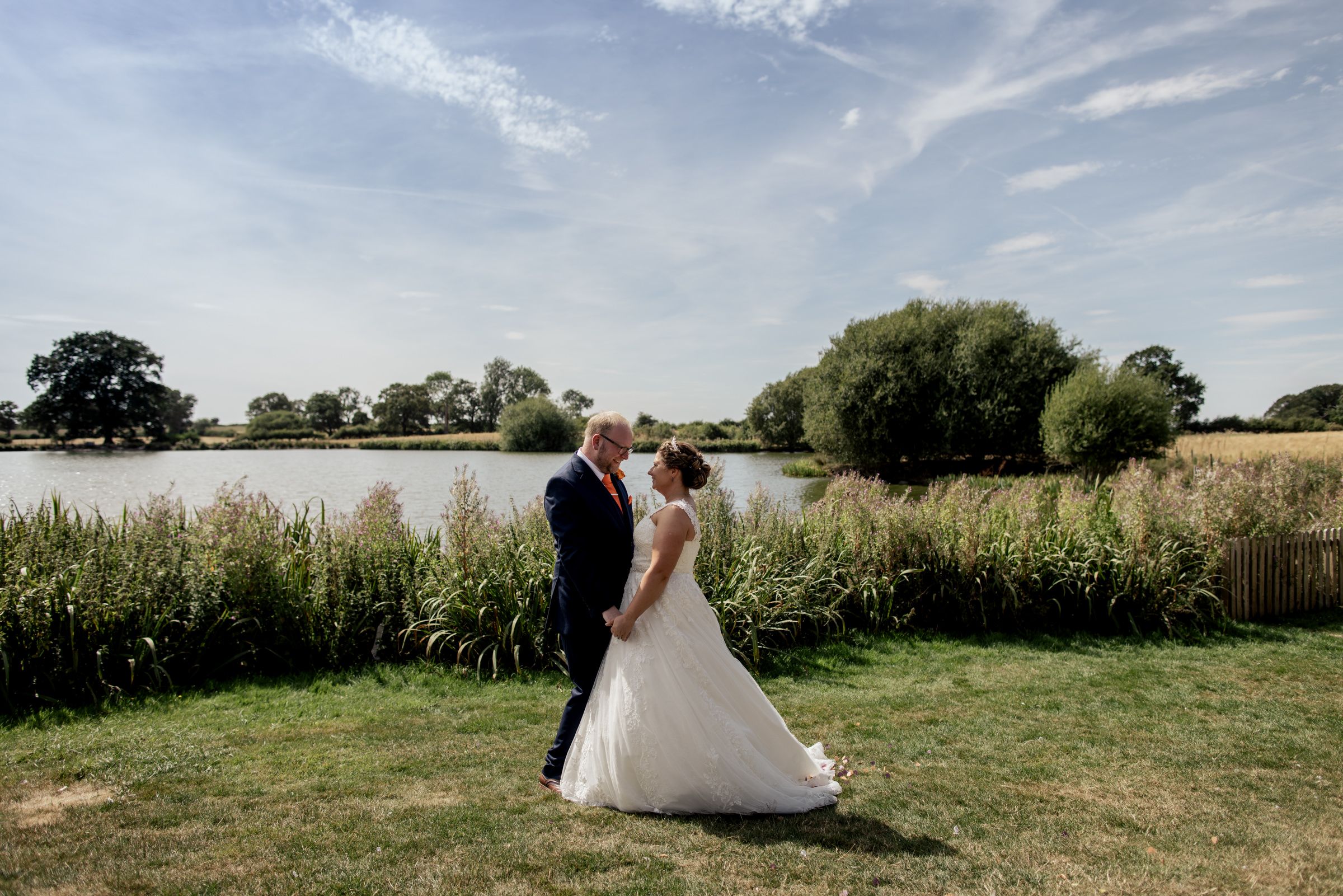 Bride and groom share a romantic moment by a serene lake surrounded by lush greenery on a sunny wedding day.