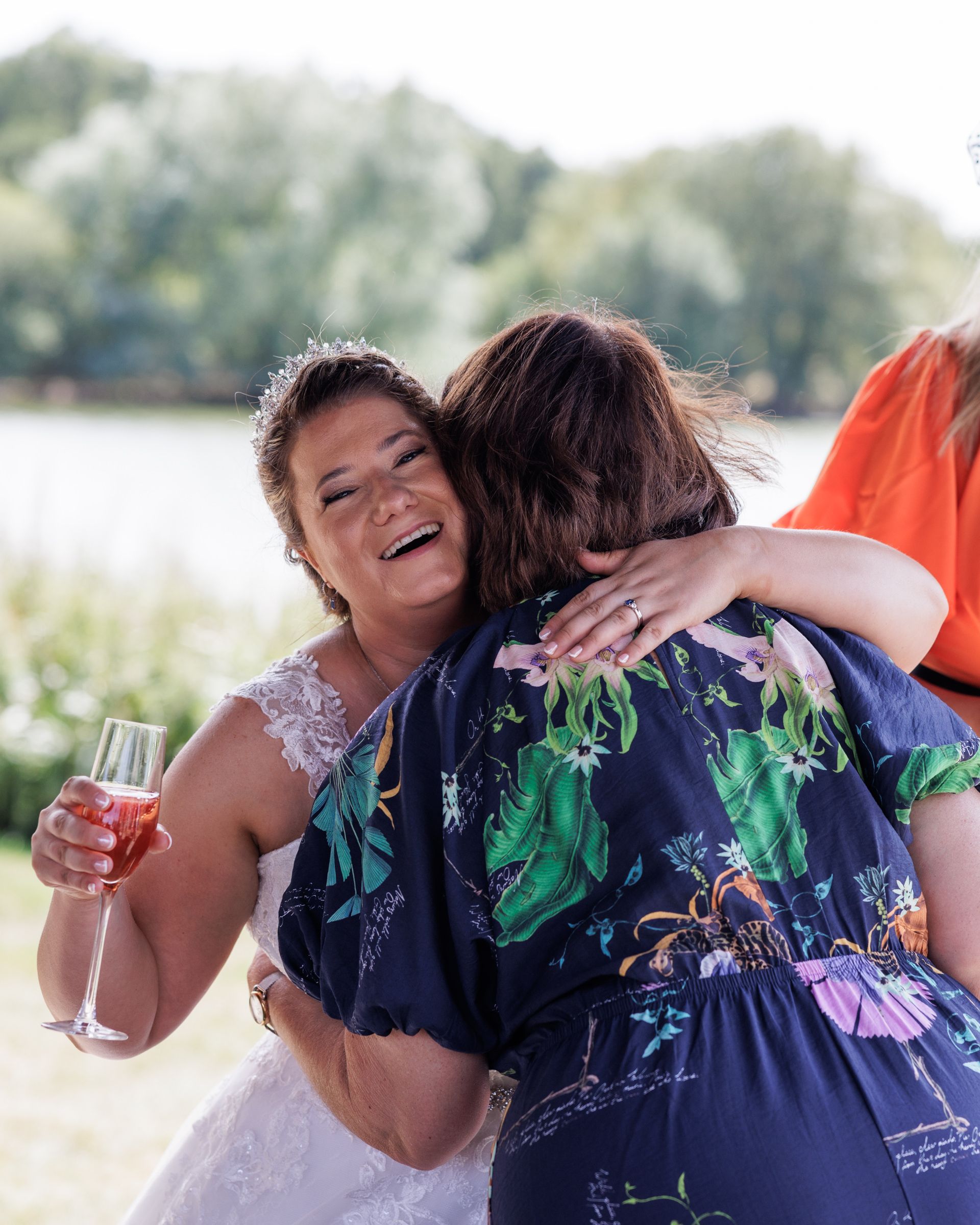 A joyful bride in a lace dress hugs a guest in a floral top while holding a pink champagne glass outdoors.