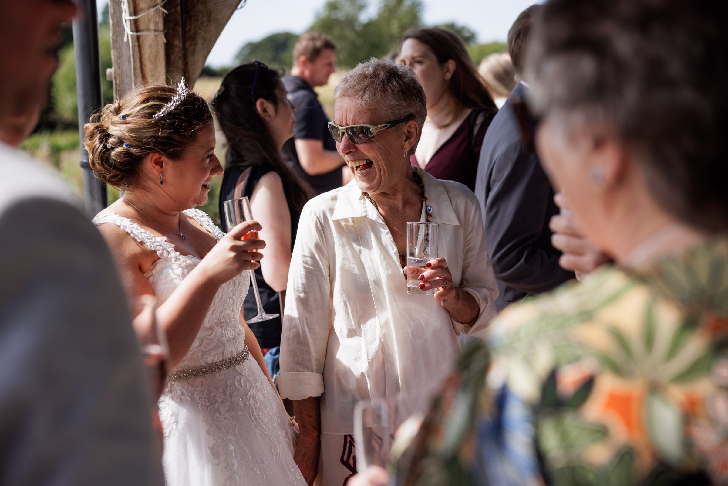 Guests socializing at an outdoor wedding reception, with a smiling woman in sunglasses holding a drink.