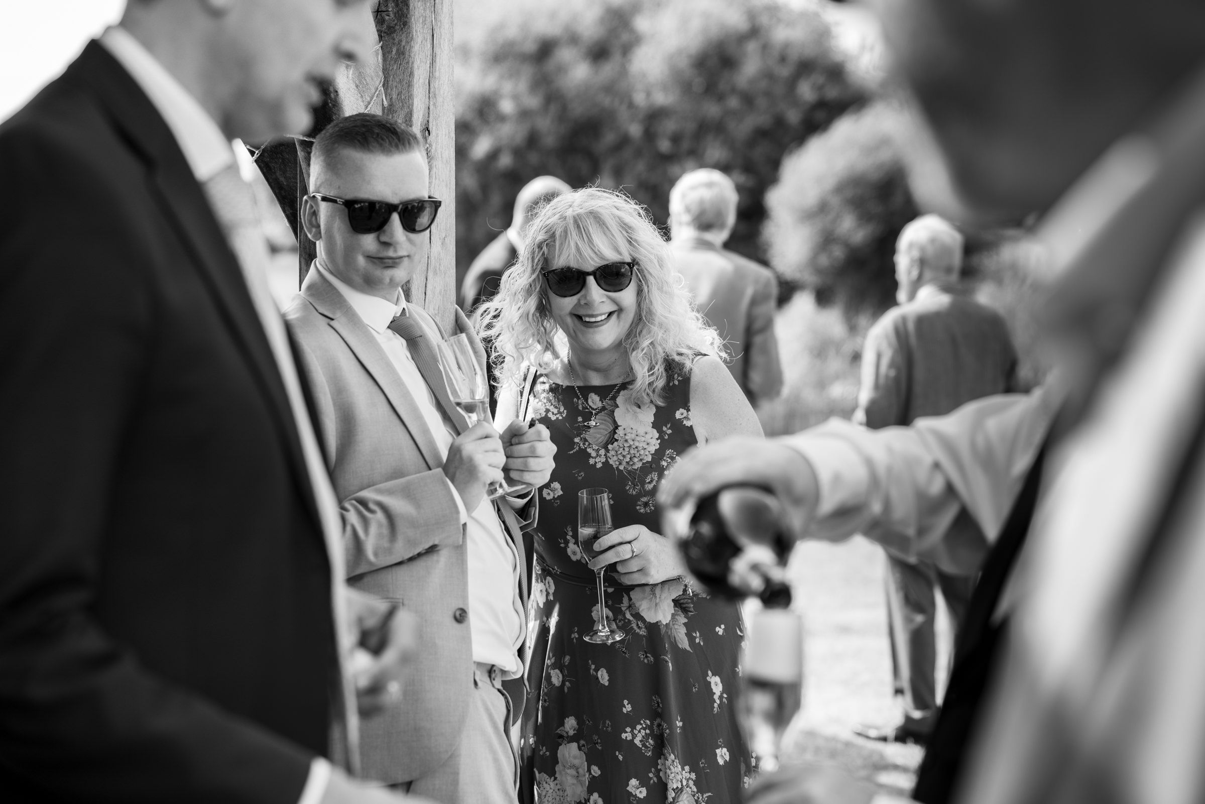 Black and white photo of wedding guests socializing outdoors, smiling woman in floral dress holding a champagne glass.