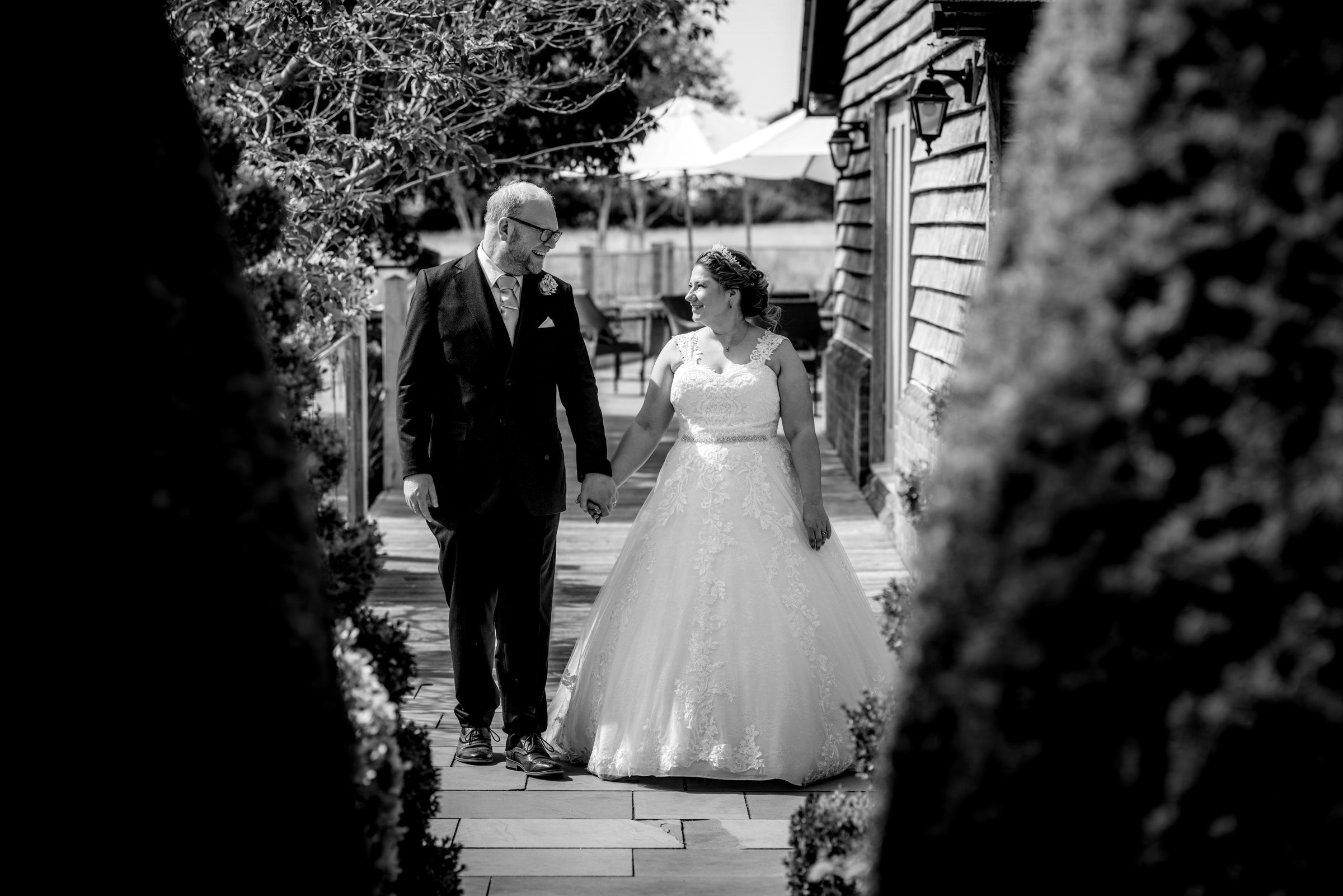 Black and white wedding photo of bride in ball gown walking with older gentleman through garden pathway.