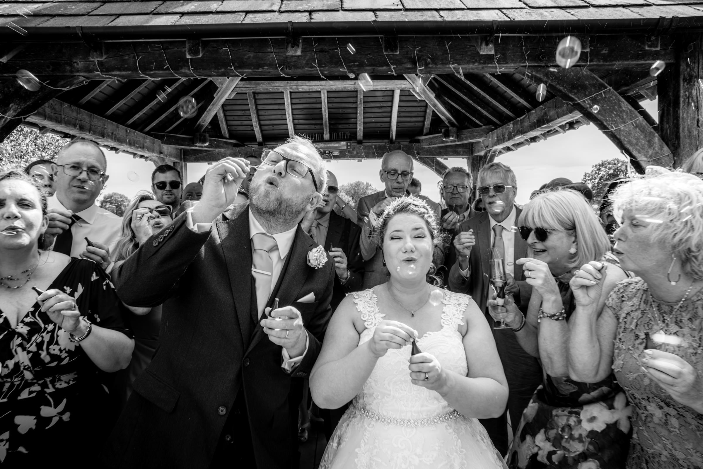 Black and white wedding photo of bride and groom blowing bubbles with guests celebrating under a gazebo.