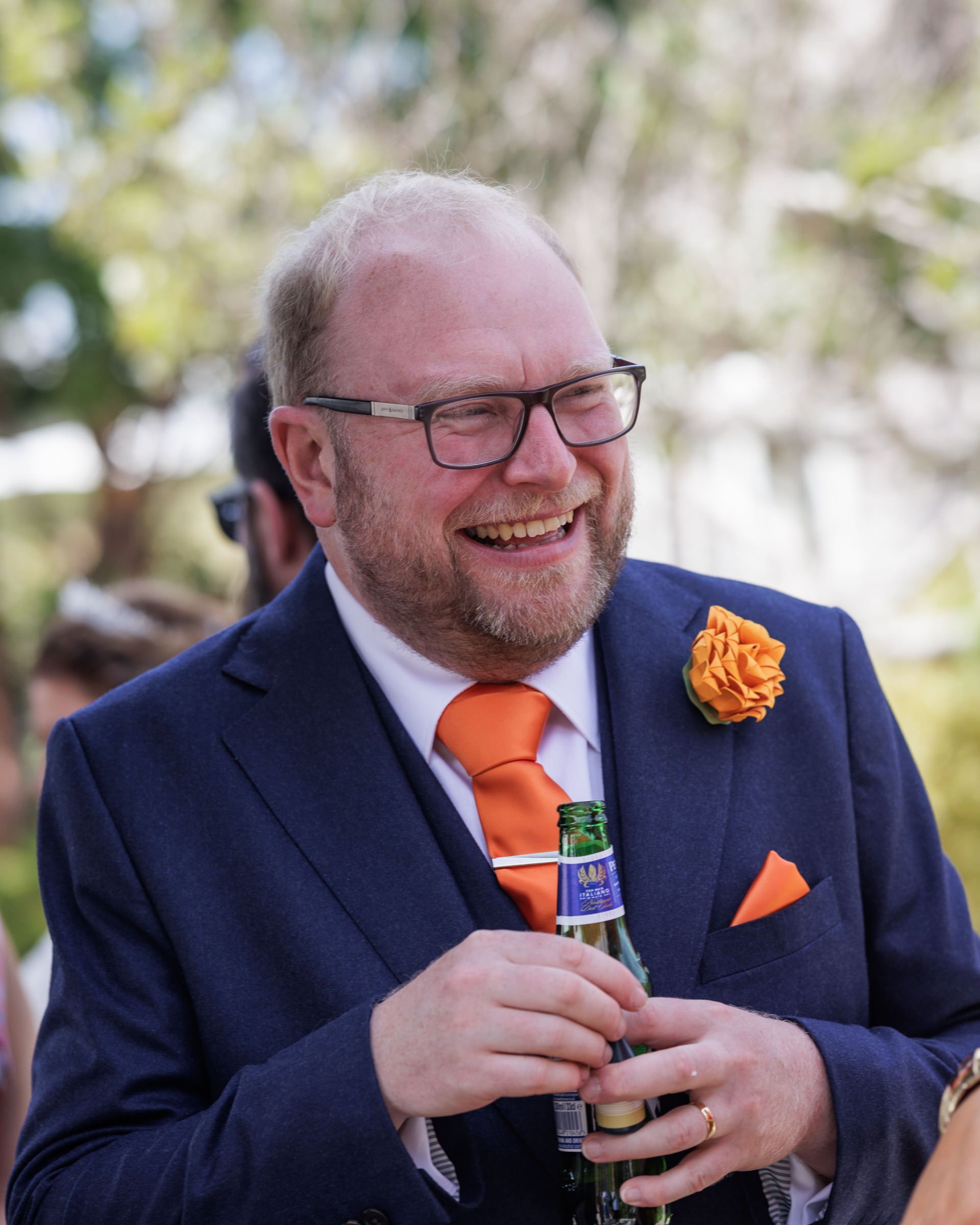 Smiling man in navy suit with orange tie, boutonniere, and pocket square holding a beer bottle outdoors.