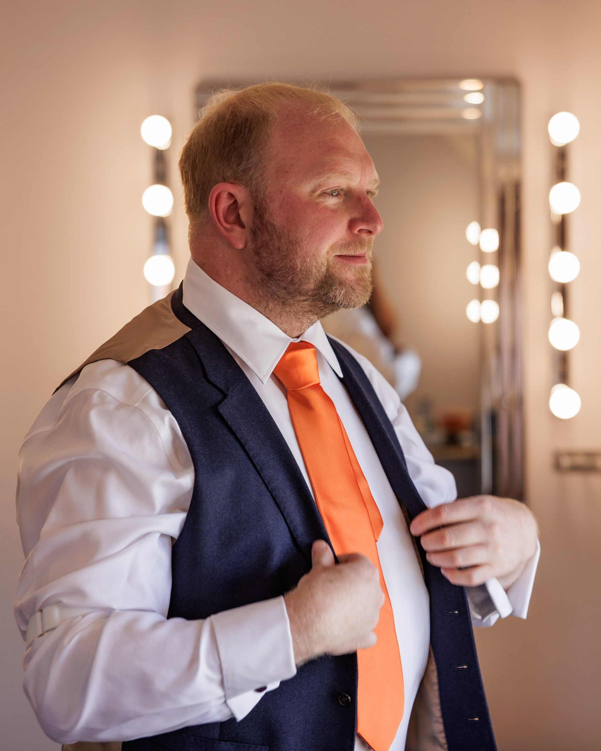 A well-dressed groom in a navy vest and orange tie adjusts his jacket in a mirror-lit dressing room.