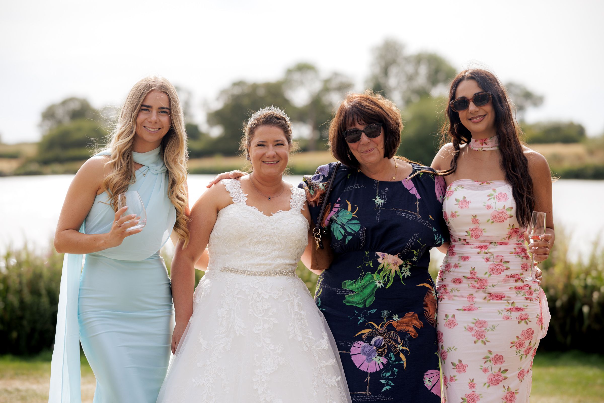 Four women posing outdoors at a wedding, one in a white bridal gown, others in elegant dresses near a lake.