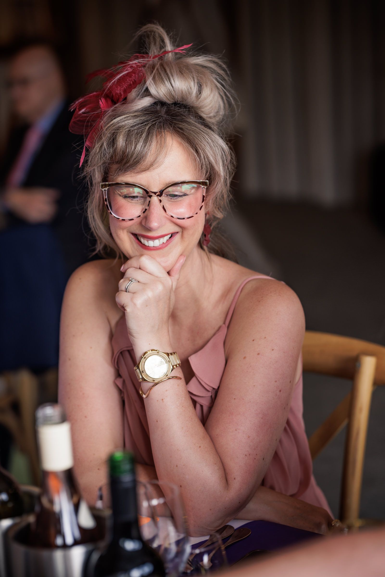 Smiling woman with updo hair, decorative glasses, gold watch, and pink dress laughing at a table with wine bottles.