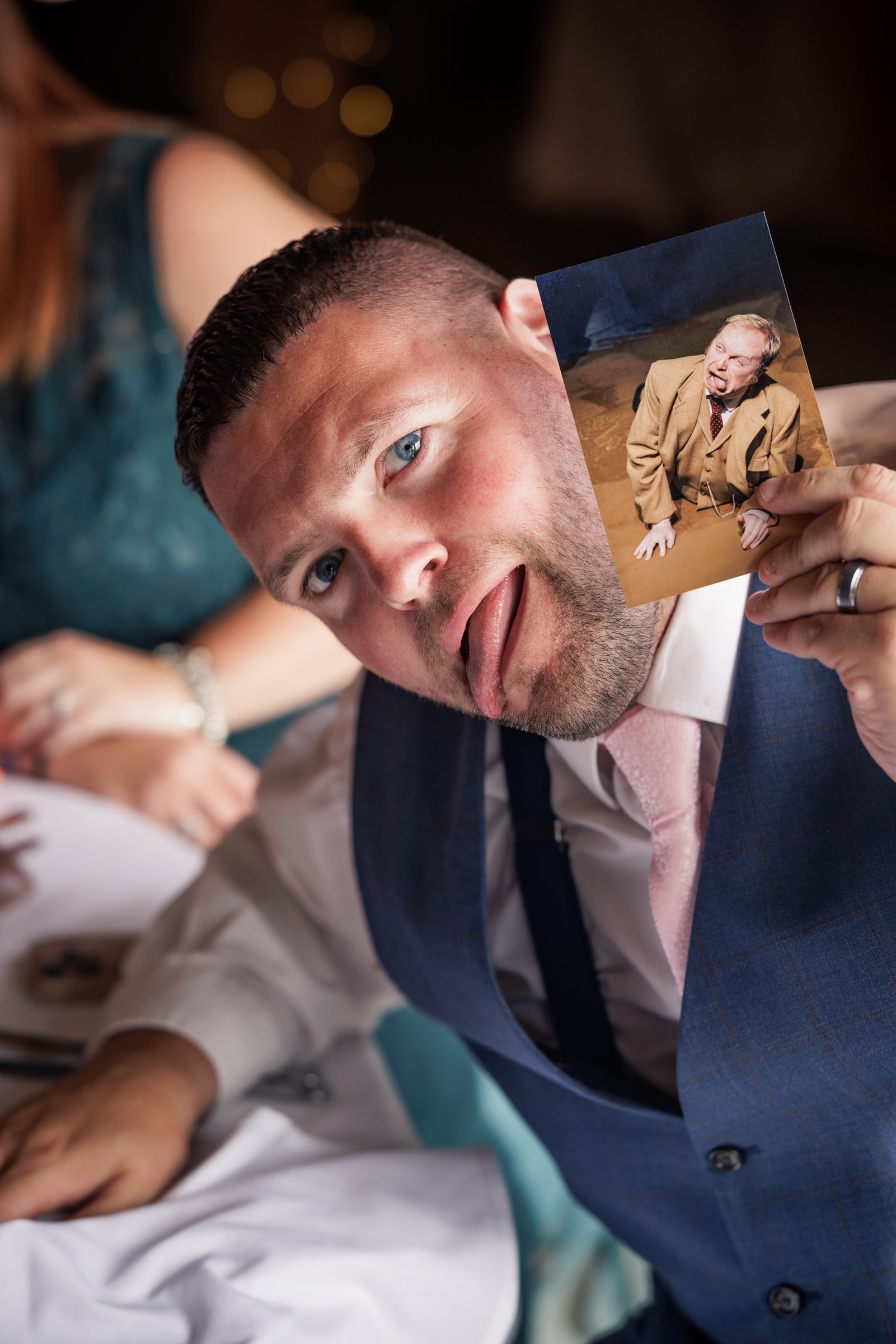 A man in a blue suit holds up a photo while making a surprised expression at a formal event.