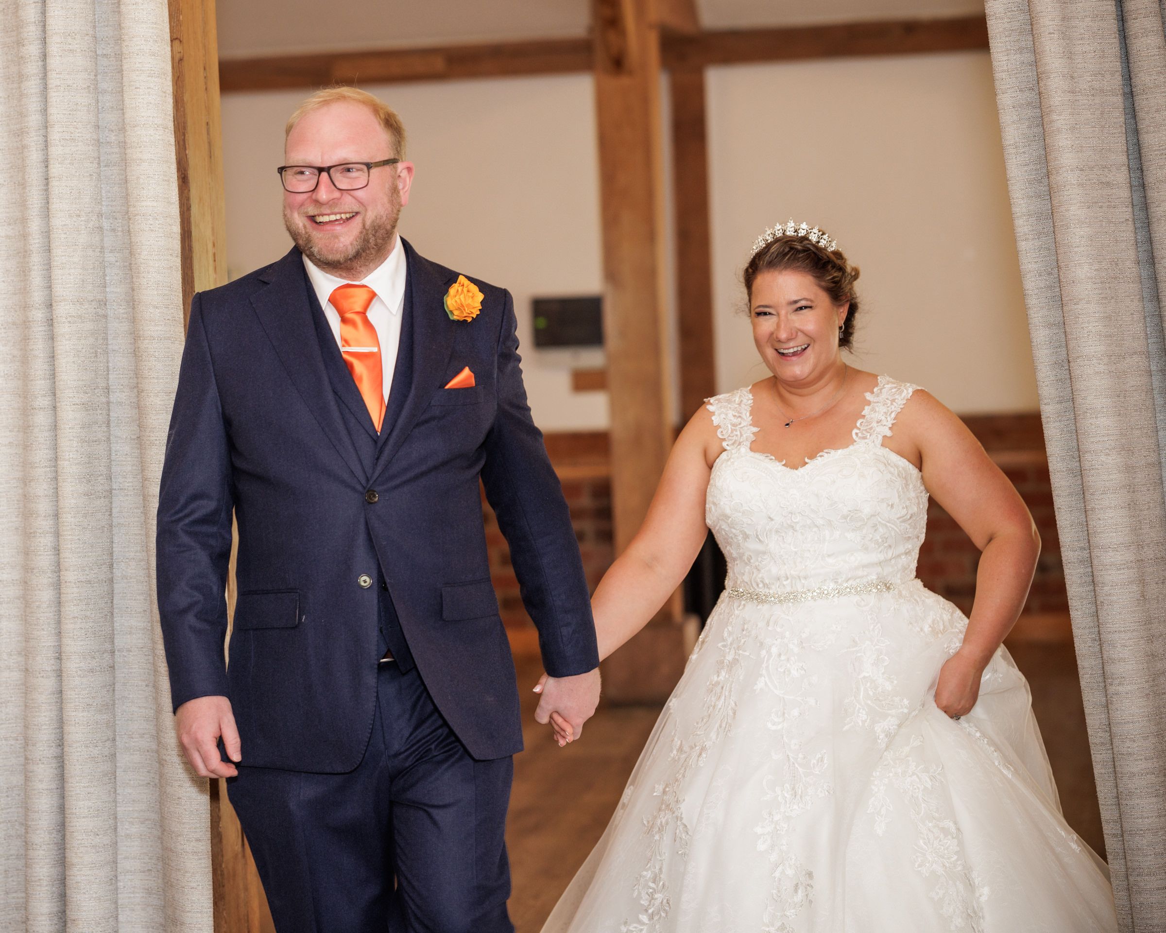 Bride and groom holding hands walking into wedding reception, groom in navy suit with orange tie, bride in white lace gown.