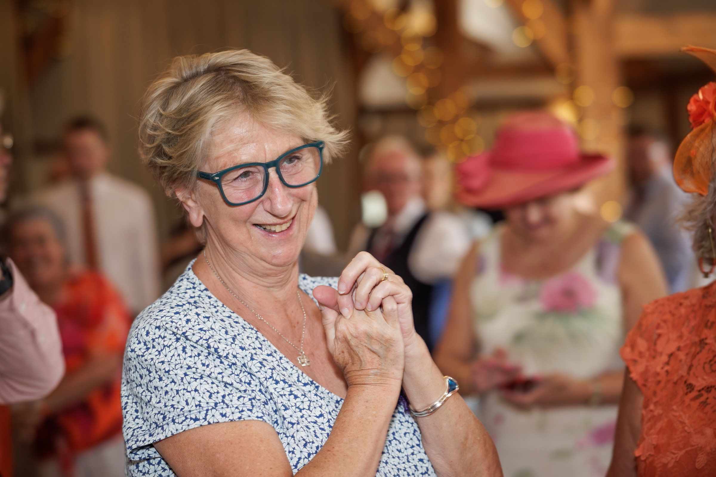 Smiling older woman with glasses in floral dress at elegant indoor social gathering with guests in colorful hats.