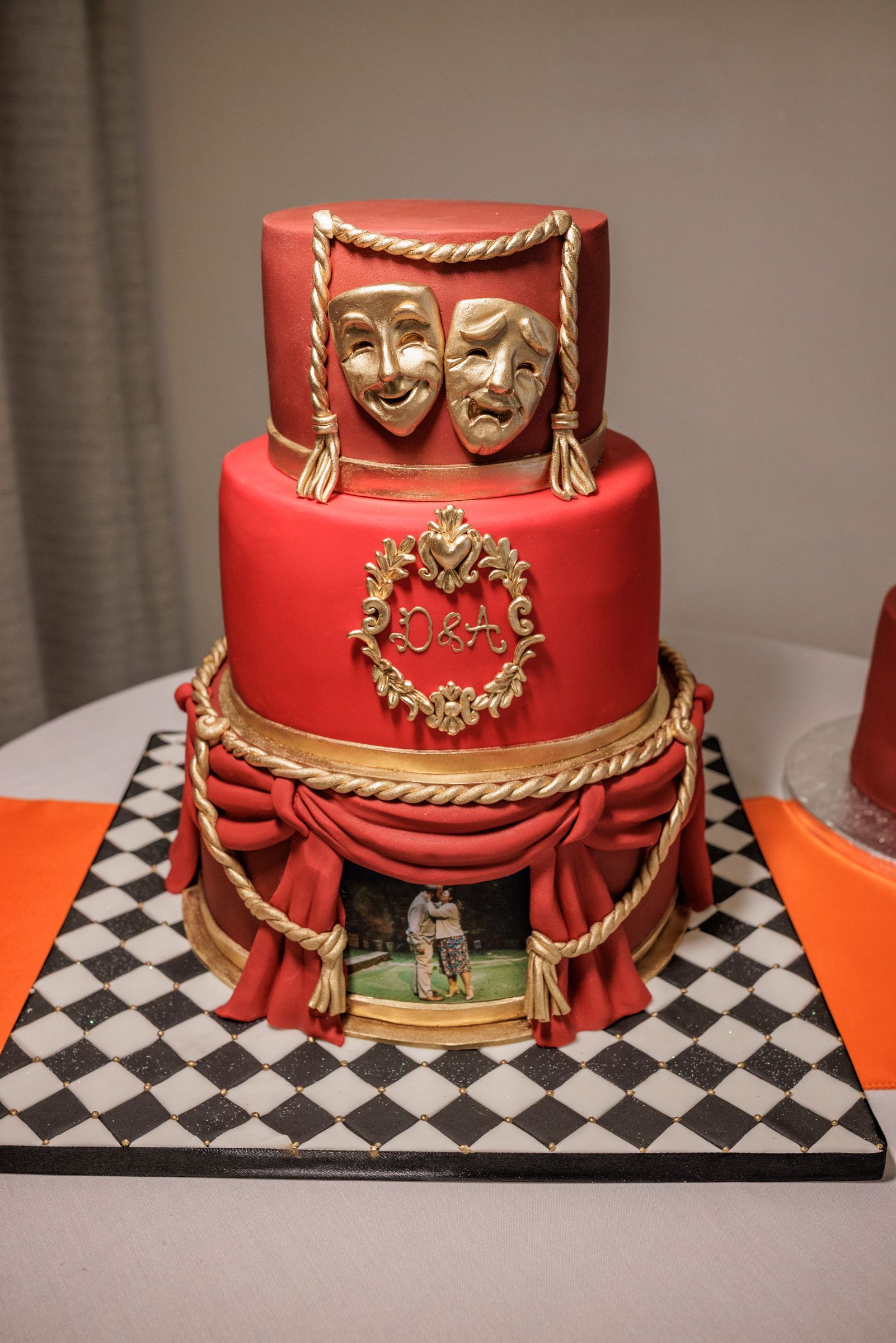 Red three-tier theater-themed cake with gold comedy tragedy masks, rope accents, and checkered base board.