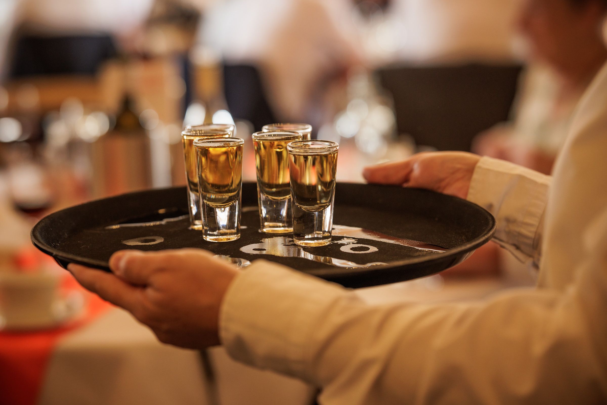 Waiter serving four shot glasses of amber liquor on a round black tray in an elegant restaurant setting.