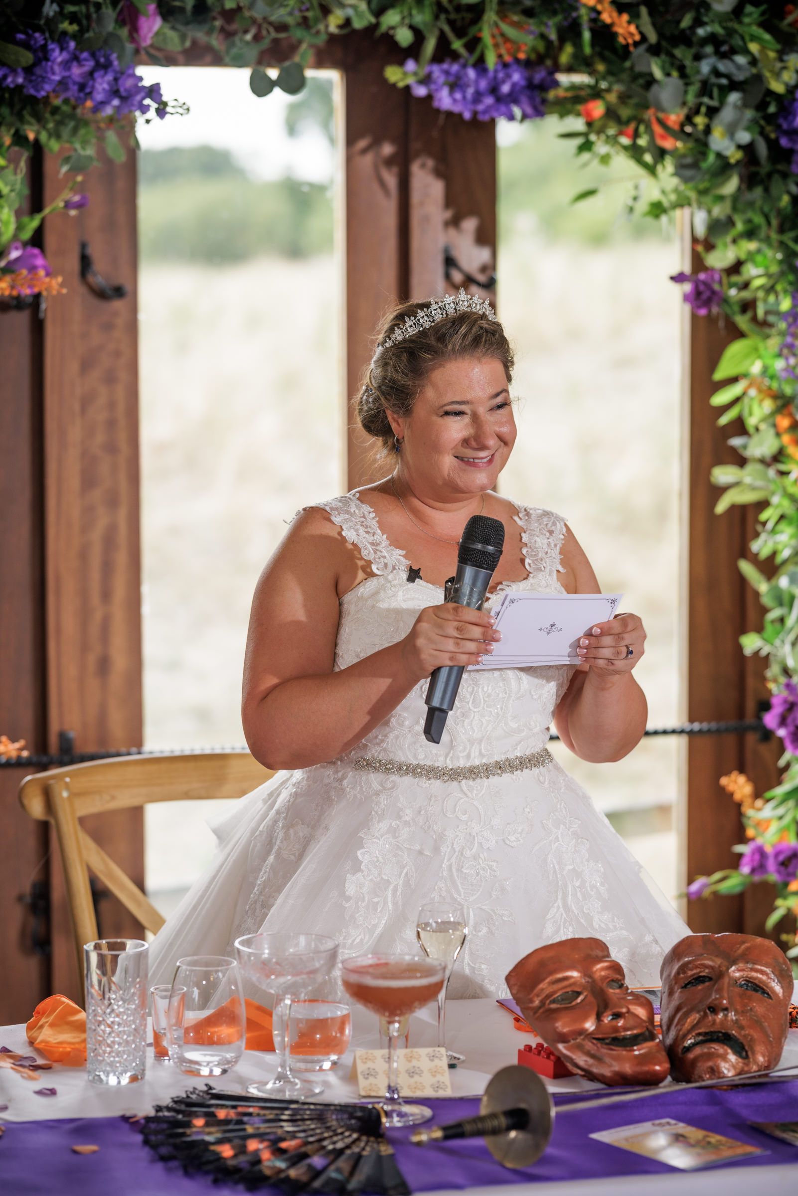 Bride in white lace gown giving speech at wedding reception with purple table decor and floral arch.