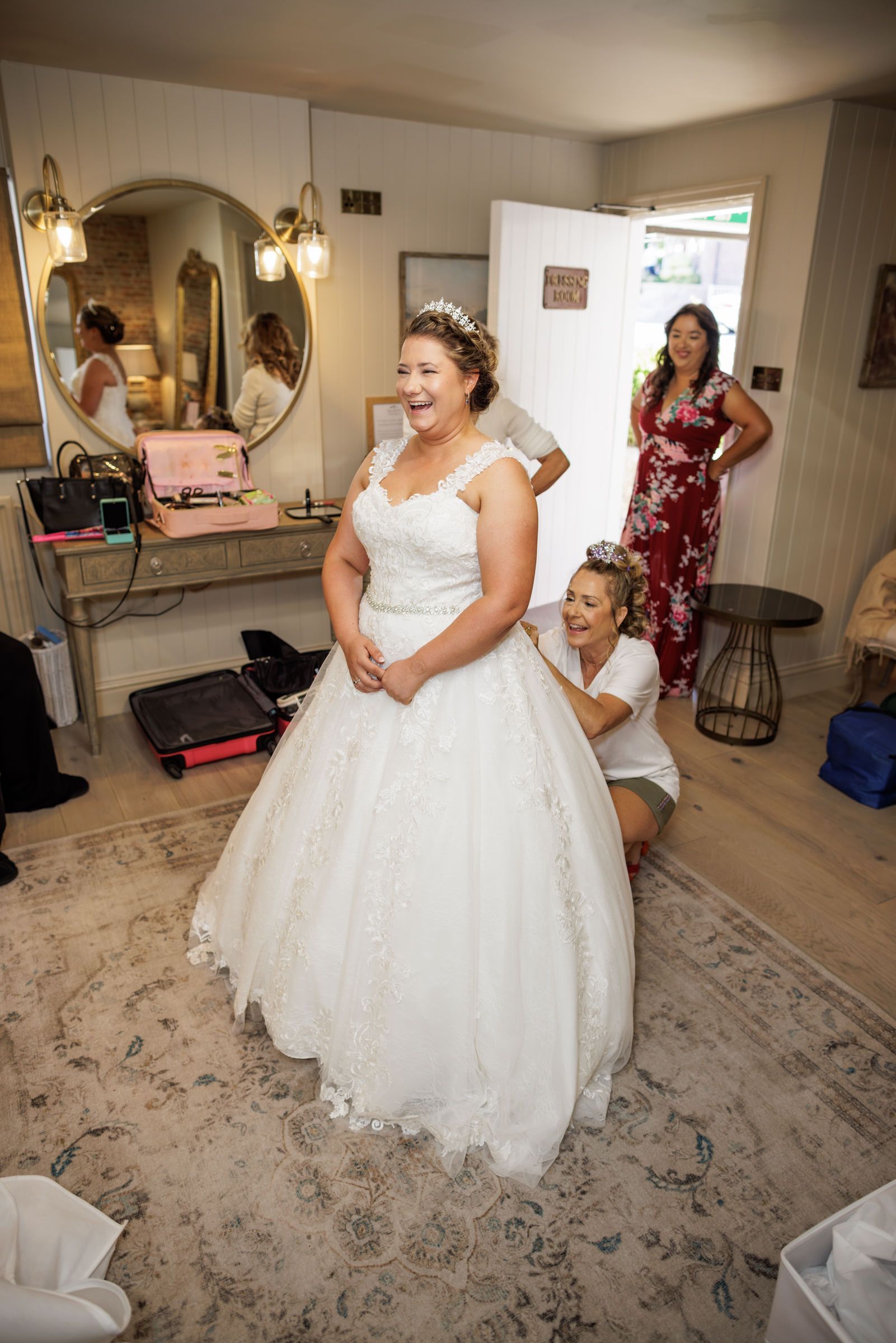 Bride laughing in white ballgown wedding dress being helped by bridesmaid while another guest watches in bridal suite.